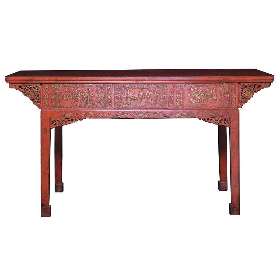 Chinese red lacquered painting table, soft wood construction with unusual detailed relief skirt carving depicting figures and flowers, three deep drawers with inset brass hardware, intricate pierced corner carvings, splayed leg design with end cross