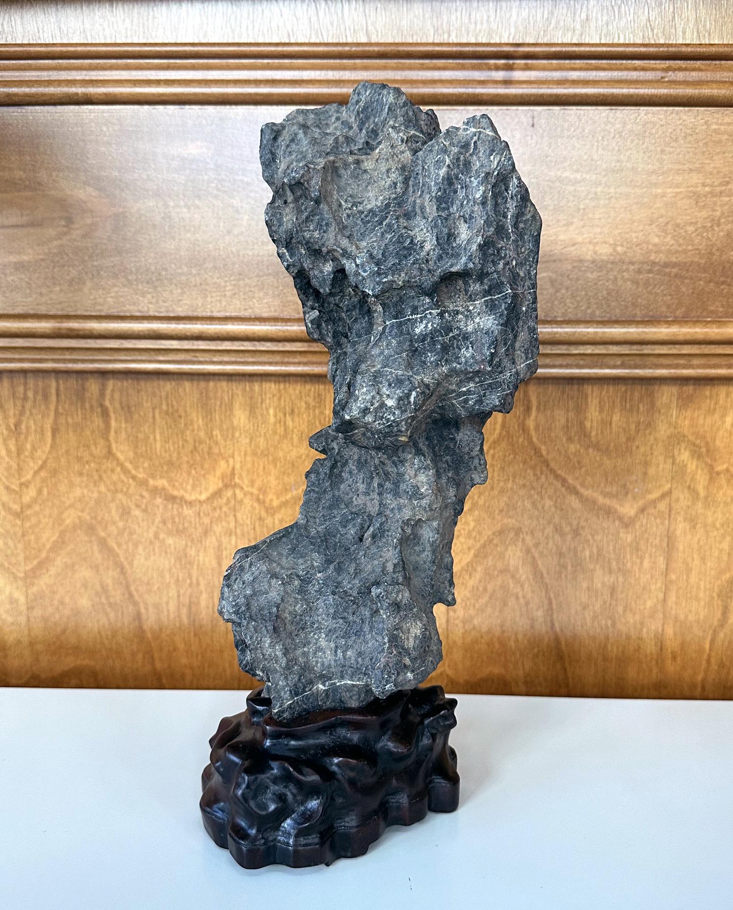 An intriguing Chinese scholar rock in vertical form presented on a custom hand-carved wood stand circa late Qing Dynasty. The greyish black stone is of Yingde type. Its upright form is poetic and nicely balanced, reminding one the limestone