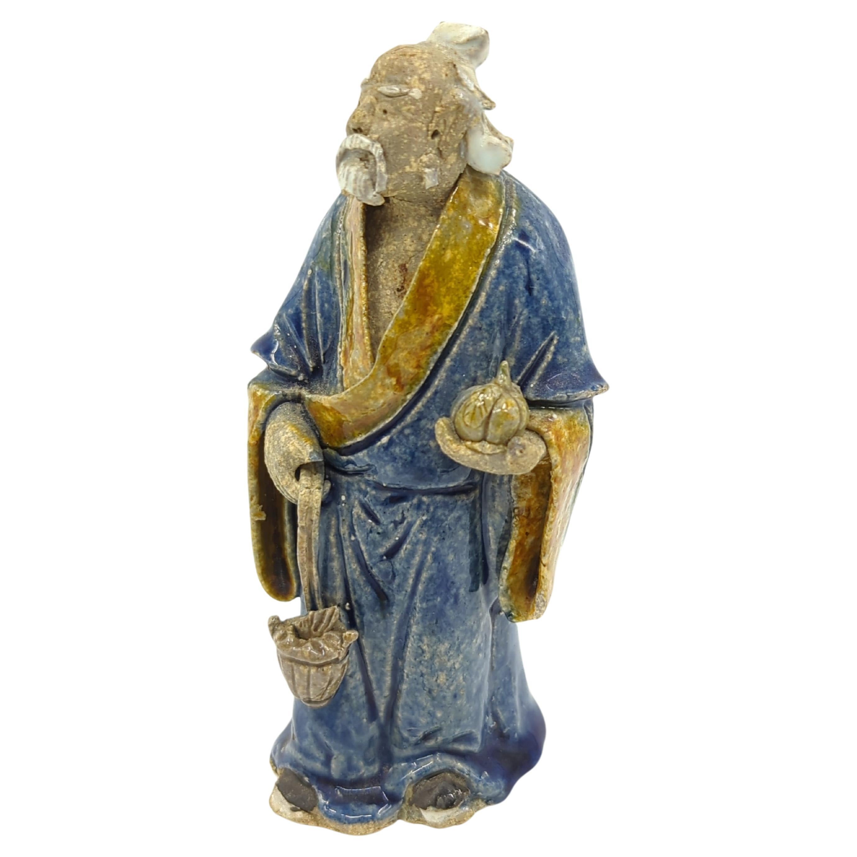 Antike chinesische, handgefertigte und handbemalte Lehmfigur eines stehenden älteren Gelehrten in blauem Gewand, der in der einen Hand einen großen, gelappten Kürbis hält und in der anderen einen gefüllten Fischkorb trägt. Die Figur hält eine