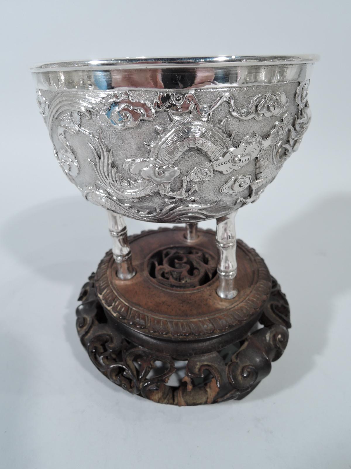 Chinese export silver bowl, circa 1900. Curved sides with chased scaly, slithering dragons on stippled ground. Circular frame (vacant). Three bamboo supports mounted to carved and bellied rosewood base. Bowl underside marked. Gross weight: 7.5 troy