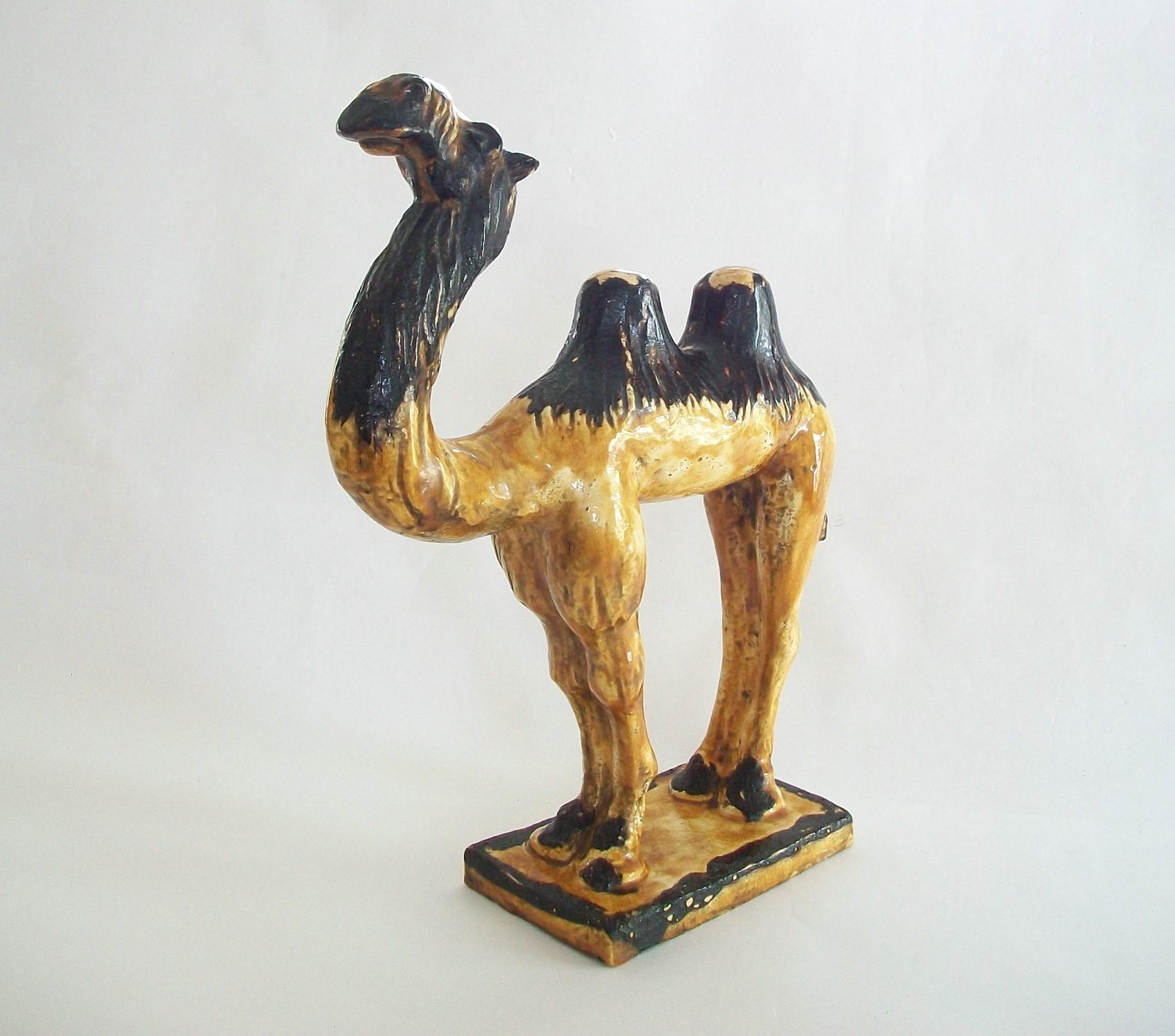 Antique Tang style Bactrian camel tomb figure - featuring an over-all amber glaze with brown highlights - stylized fur carved details to the head, legs and humps - unsigned - China - faded old collectors label fixed to the base with typewritten