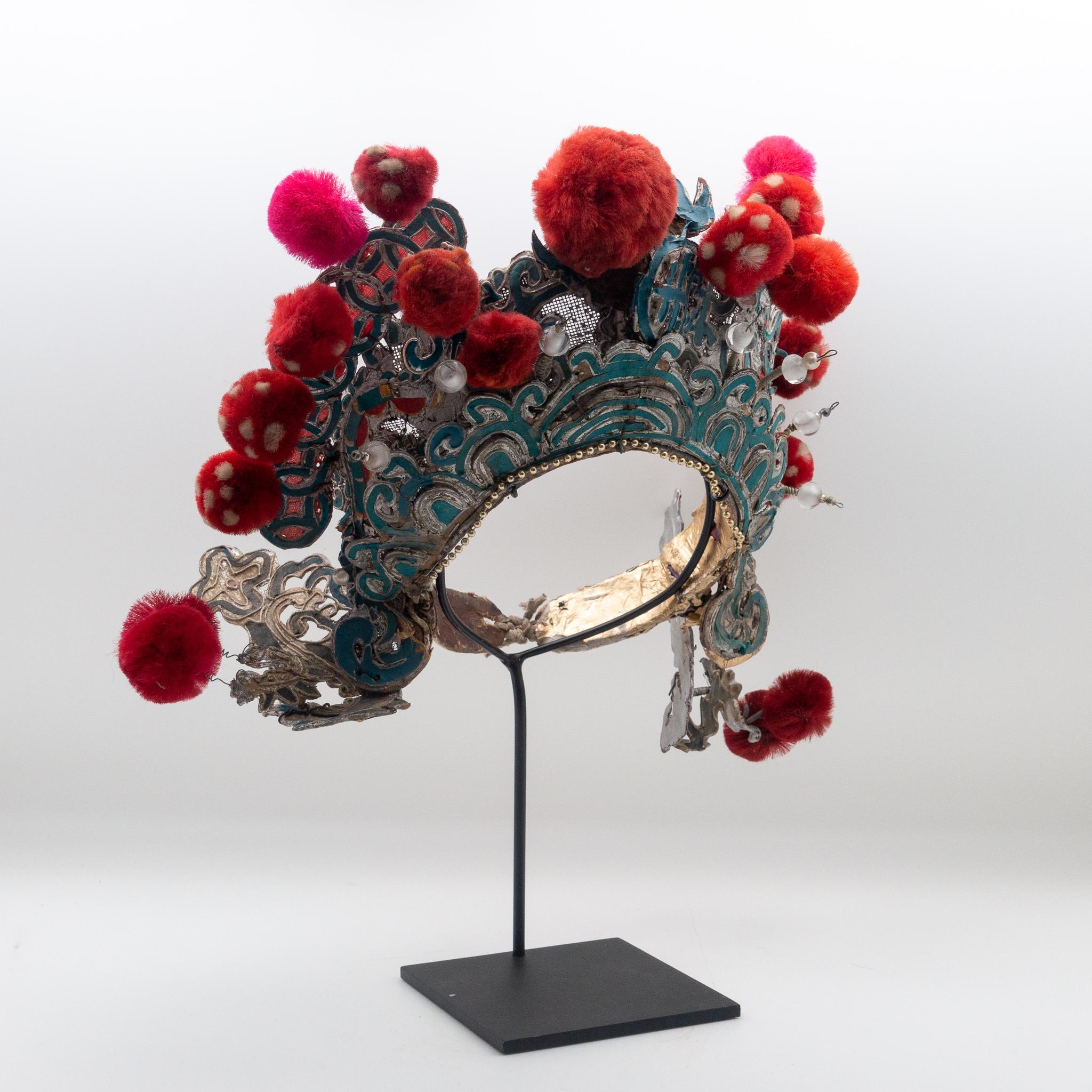 Chinese opera theatre headdress in turquoise and silver colors with red and fuchsia colored pom poms along with faux pearls, early 20th century, mounted on a custom black painted metal base.