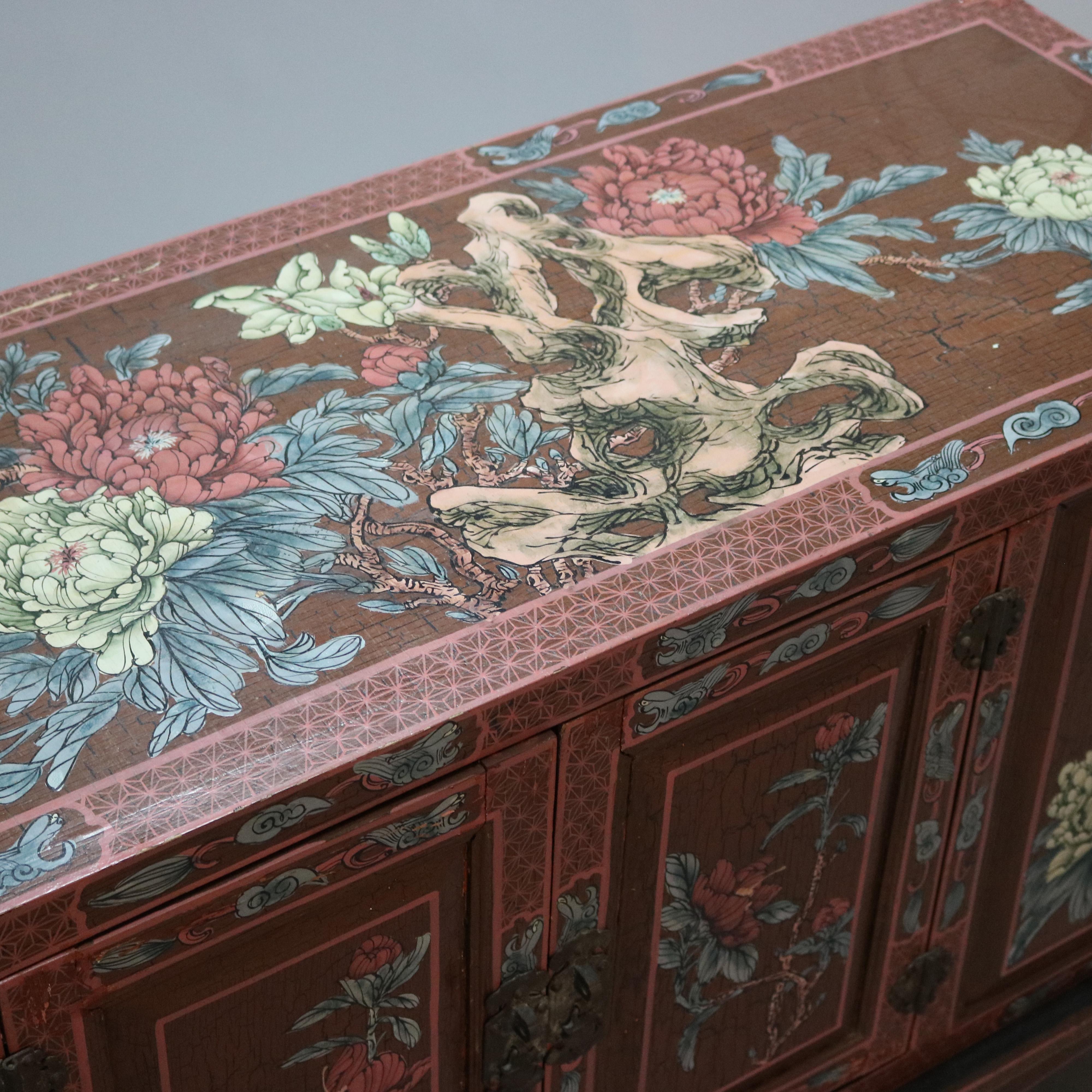 An antique Chinoiserie credenza offers wood paneled case with decorated exterior having hand painted garden flowers and two doors opening to ebonized interior, raised on matching paint decorated base with stylized scroll feet, 20th