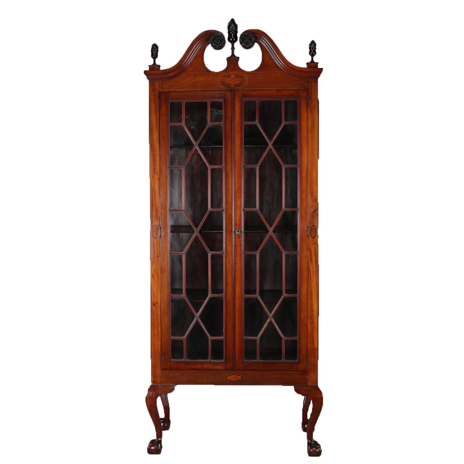 An antique Chippendale style china display cabinet featuring mahogany construction with scrolled broken arch pediment having central and flanking flame urn finials and scroll rosettes over satinwood inlaid central urn surmounting case with double