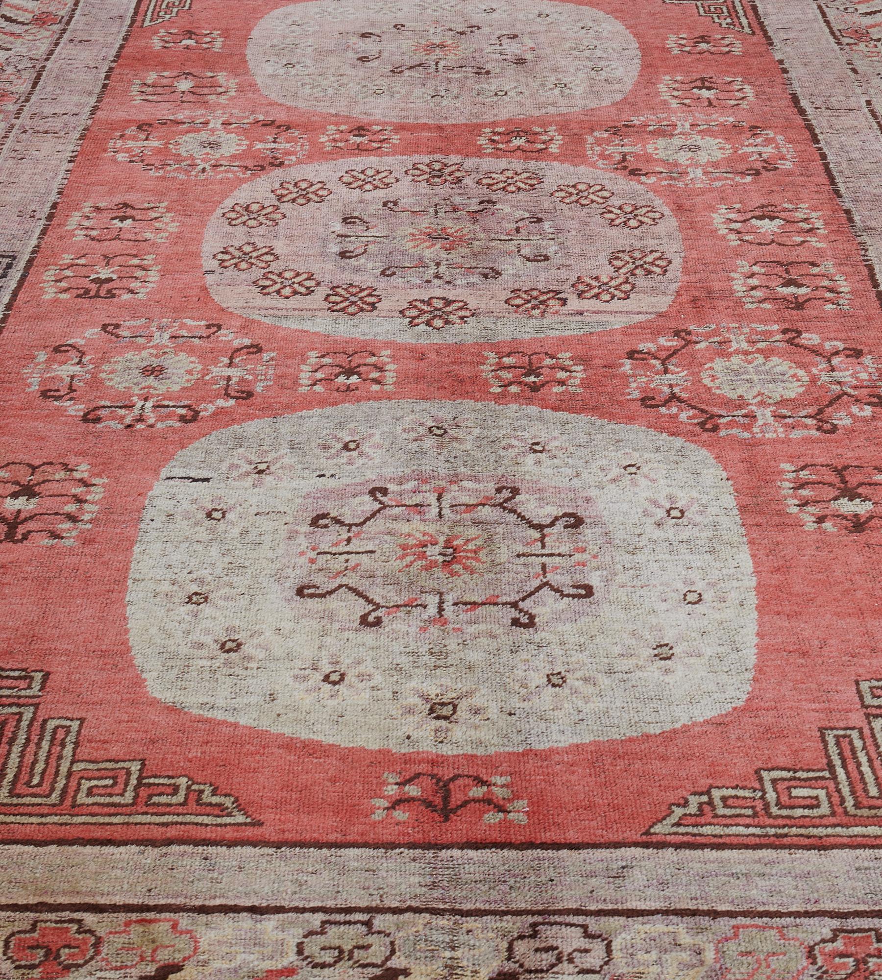 Dieser antike Khotan-Teppich aus der Zeit um 1870 hat ein korallenrotes Feld mit drei großen, schattierten, rosafarbenen Rondellen, von denen jede ein zentrales Rondell mit einem Blumenkopf enthält, aus dem eine eckige Blumenranke hervorgeht, ein