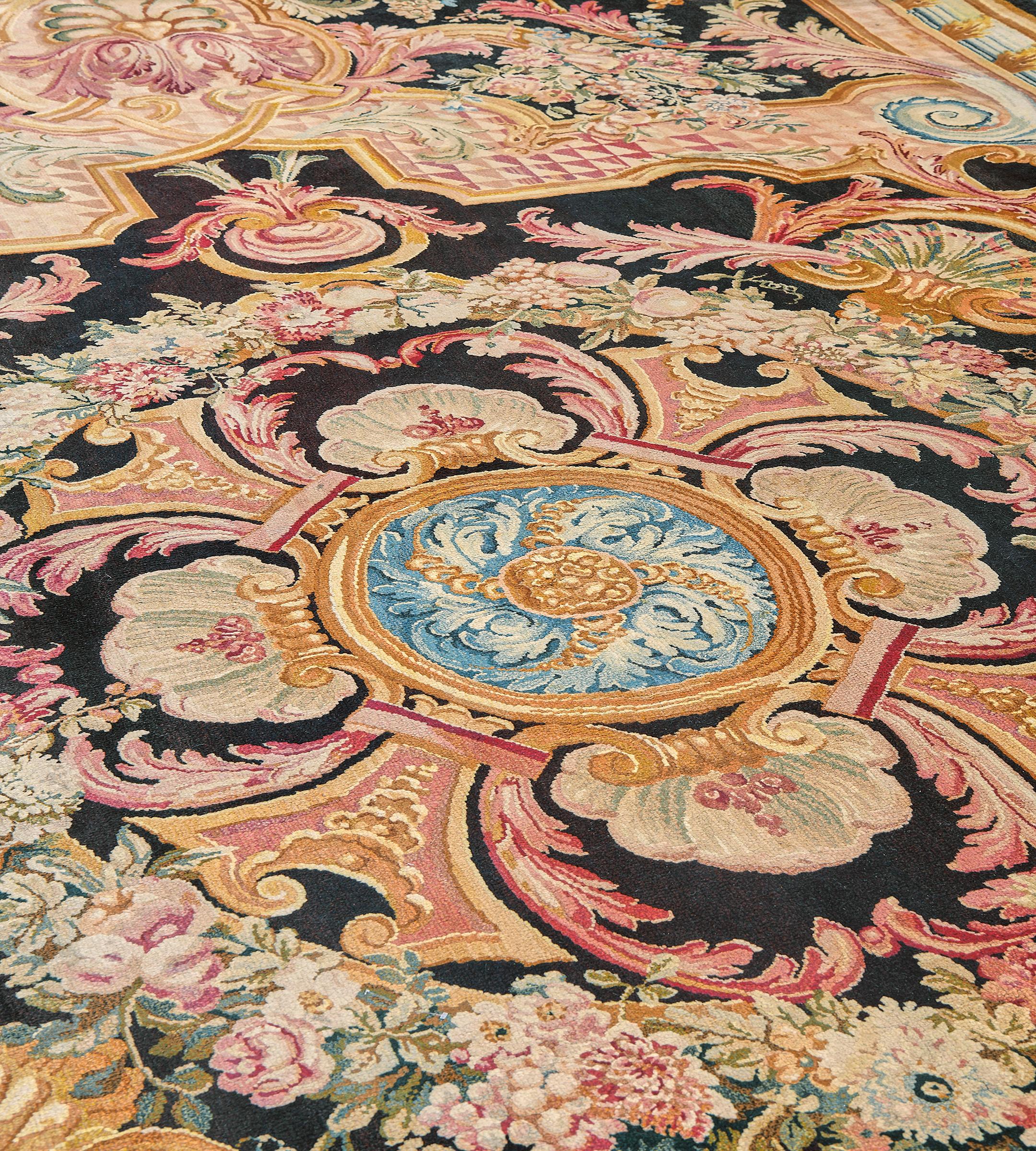 This antique, circa 1880, French Savonnerie rug features a charcoal-black field with a central panel surrounded by a bold shaded pink scrolling acanthus leaf vine forming cartouches which enclose fan-shaped shells enclosing a polychrome circular
