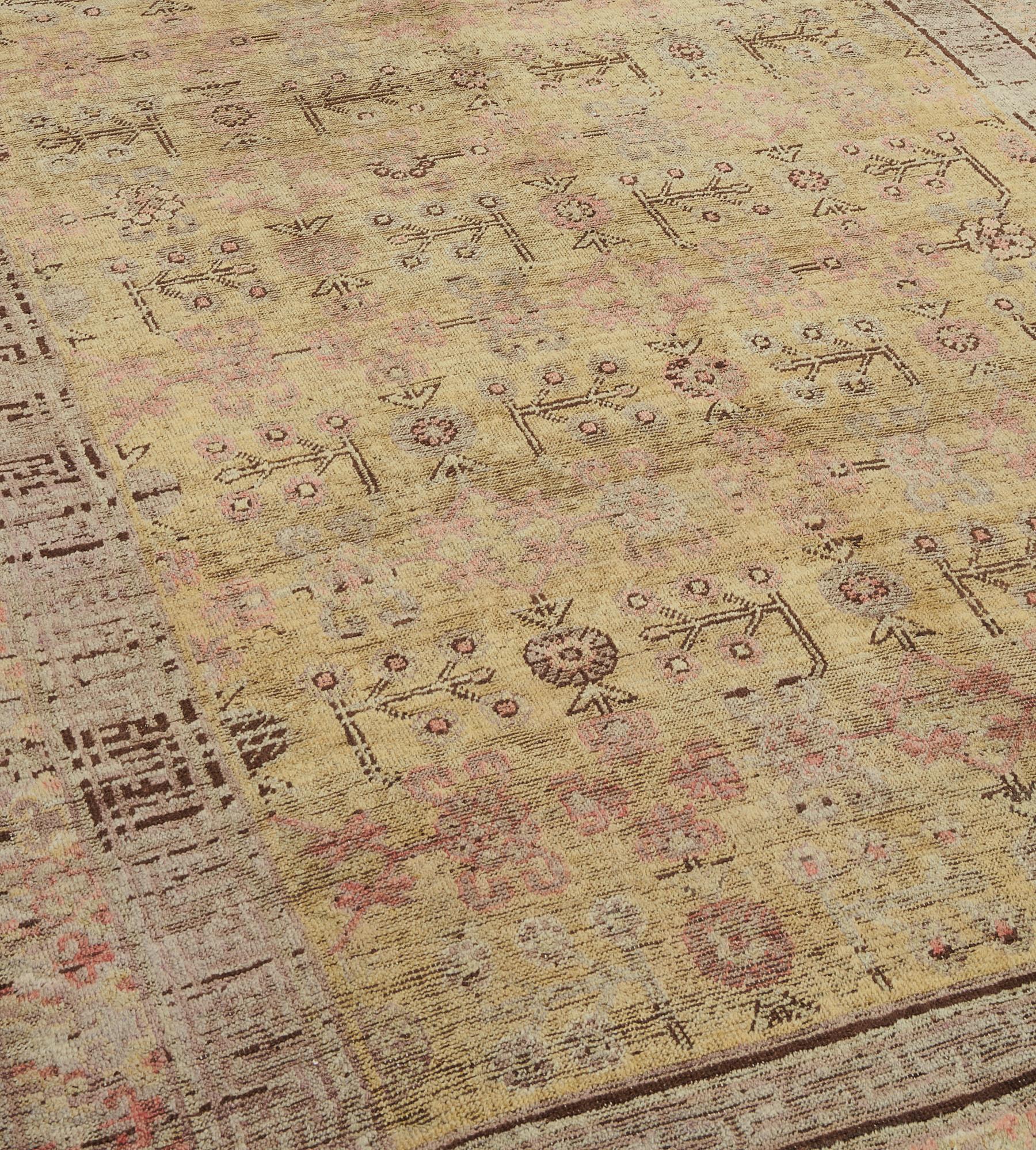 This antique, circa 1880, Khotan rug has a golden-yellow field with an overall design of delicate fox-brown angular floral stems and shaded pink flowerheads, an inner chocolate-brown and shaded ivory key-pattern square panelled stripe and an outer