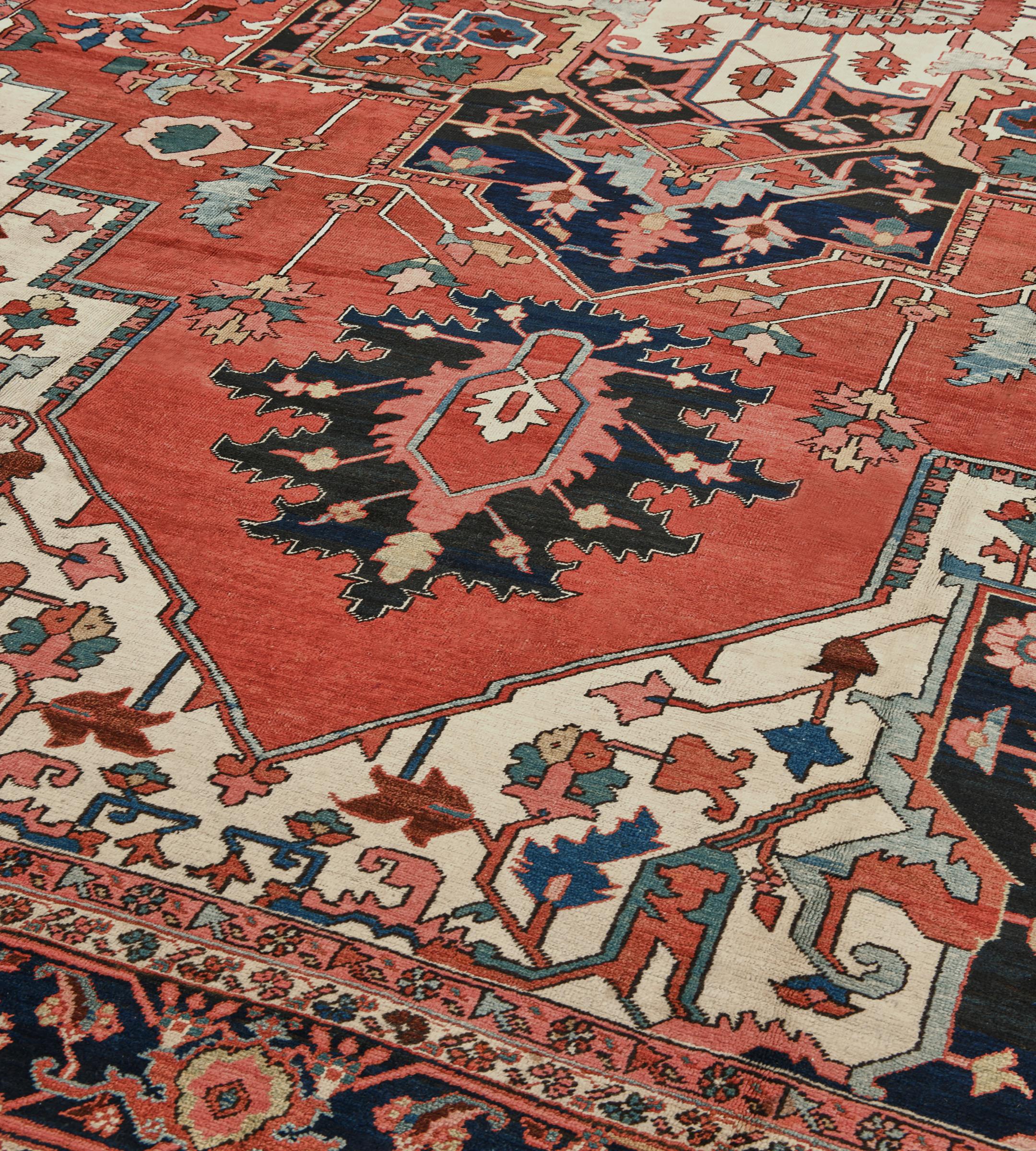 Esta antigua alfombra persa Serapi, de hacia 1900, tiene un campo rojo tomate con una enredadera angular de flores y hojas alrededor de un gran medallón central azul añil con colgantes de palmeta, con un medallón central cuspídeo de marfil con un