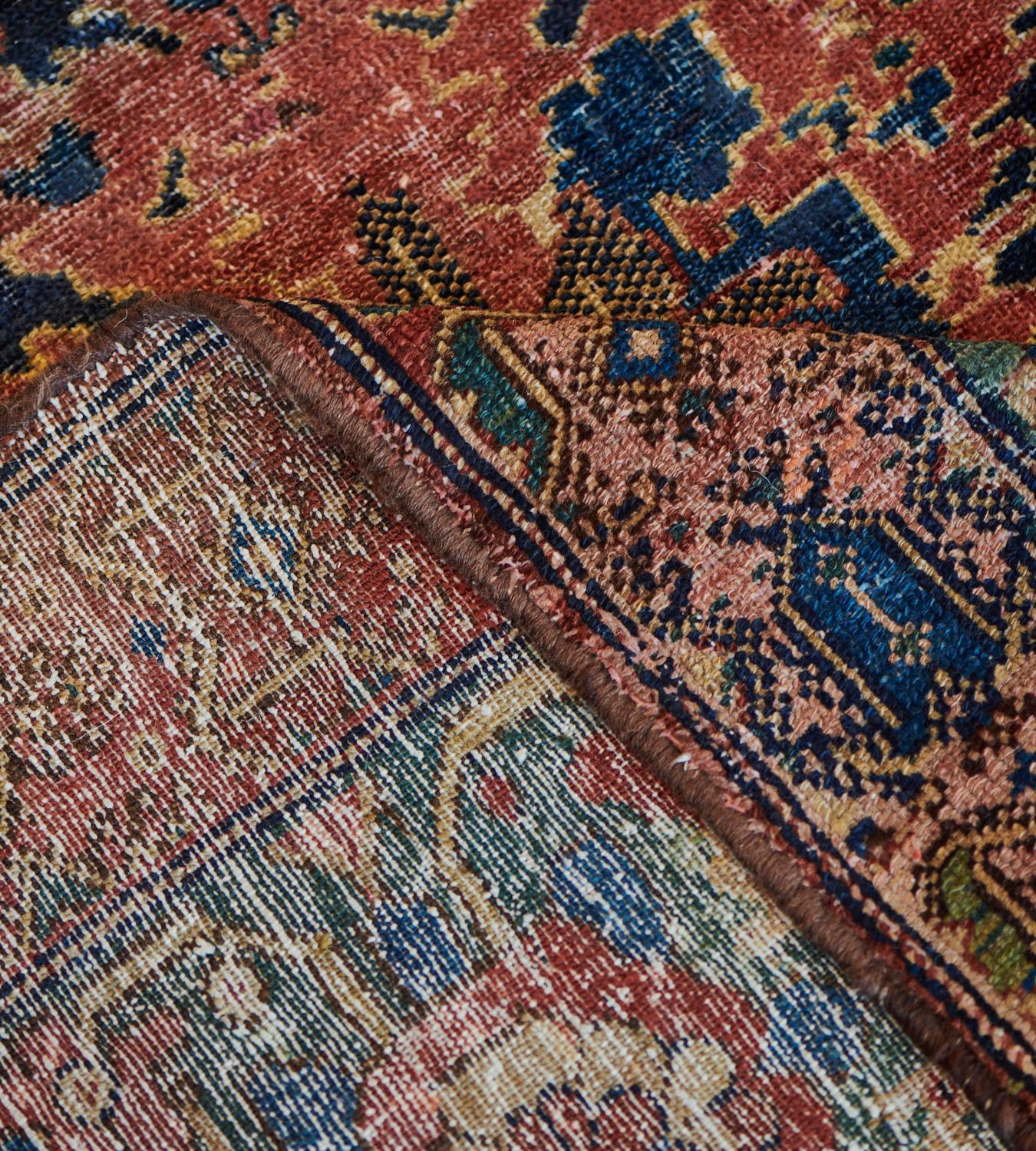 This antique, circa 1900, Malayer rug has an indigo-blue field with two broad horizontal bands of shaded terracotta-red of bold floral spays surrounded by further floral and leafy arrangements all linked by a dense floral and leafy vine, in a broad