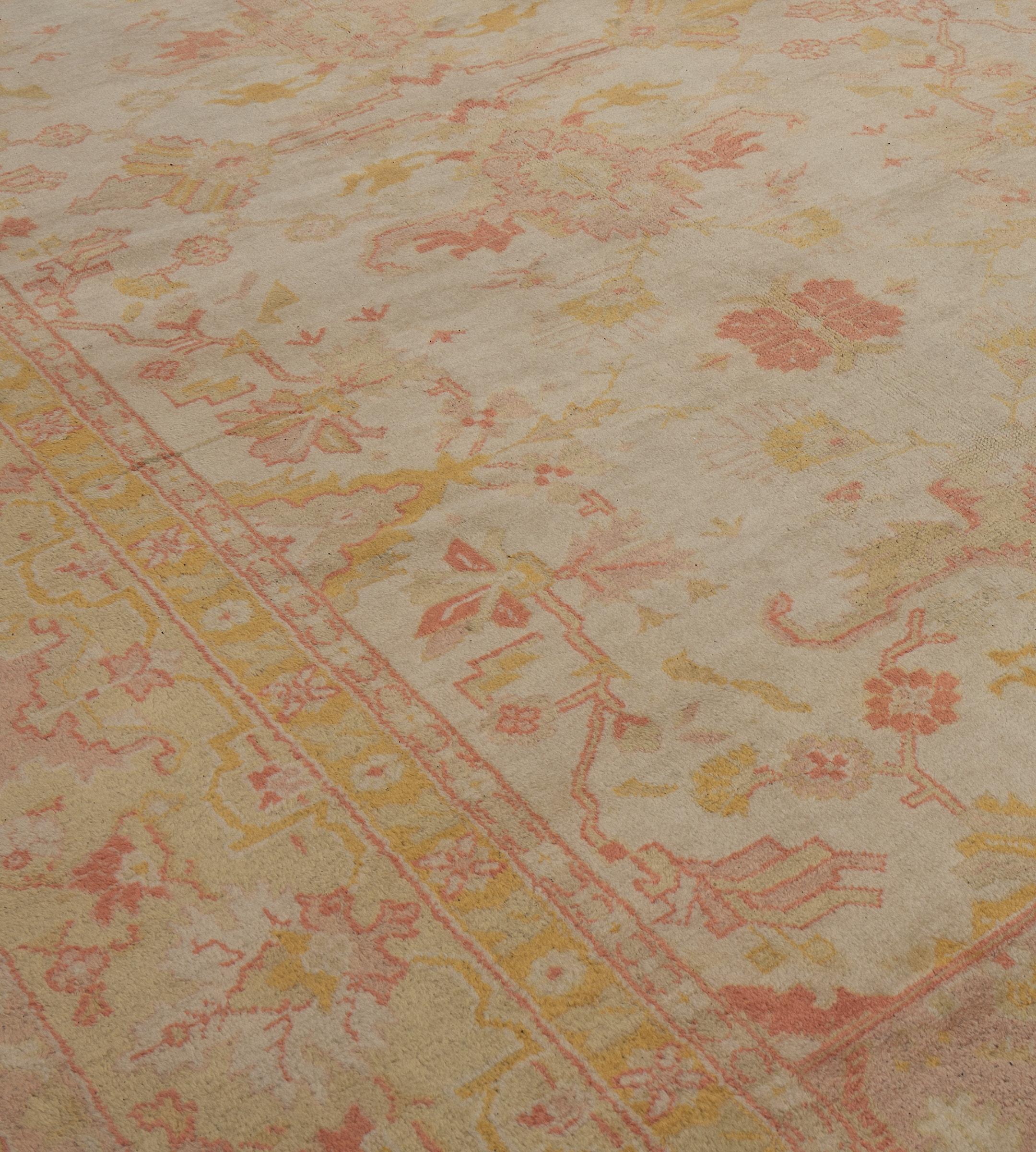Questo antico tappeto Oushak, risalente al 1900 circa, ha un campo avorio con una palmetta verde chiaro, rosa polveroso, rosso terracotta e giallo oro e un motivo floreale che collega losanghe a gradini e spruzzi floreali, un'ampia fascia rosa