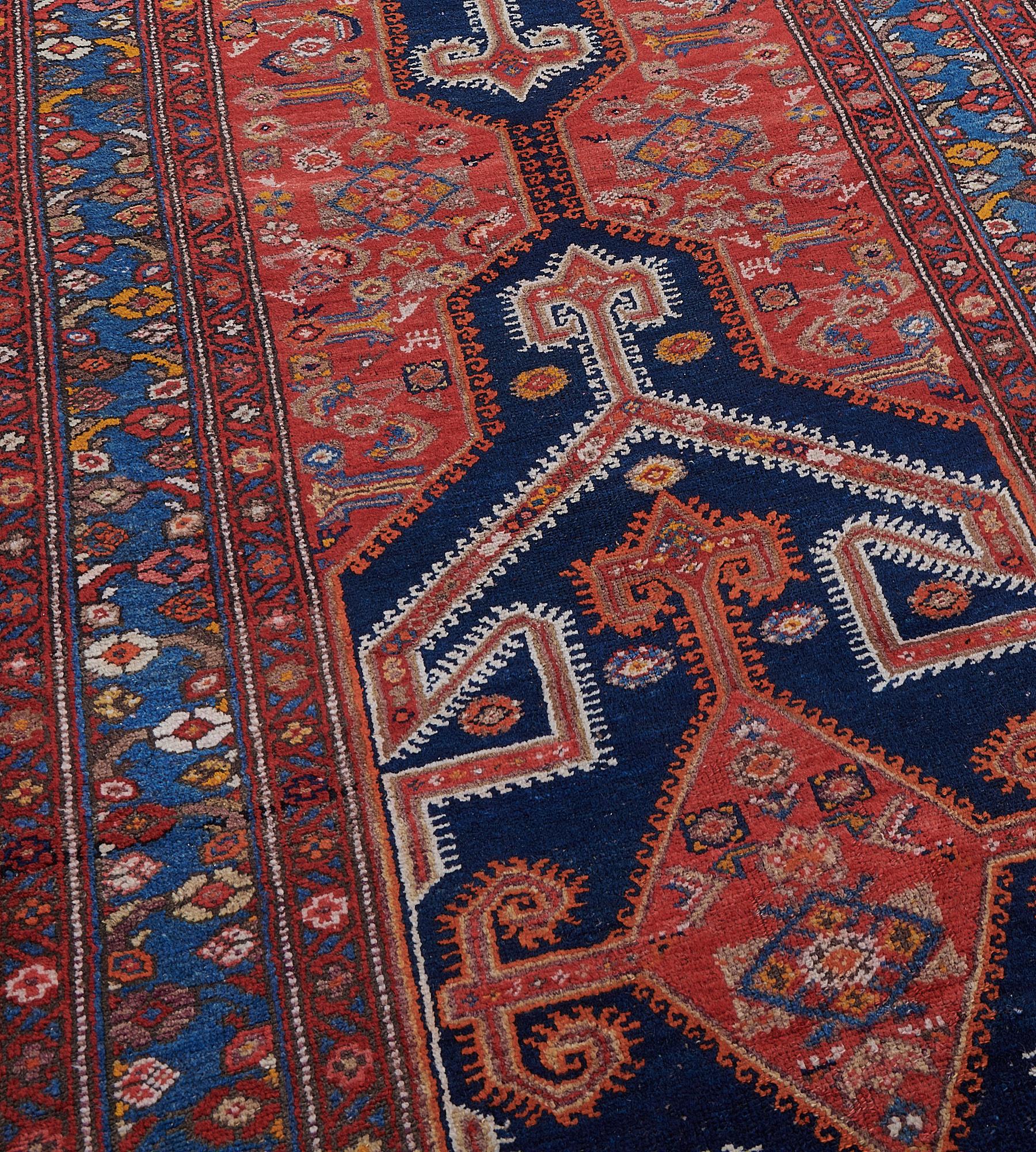 This circa 1910 Bidjar Runner has a tomato-red field with a polychrome herati-pattern around a column of three indigo-blue hooked panels each containing a central brick-red lozenge medallion with hooked motifs at each end and side an outer band of