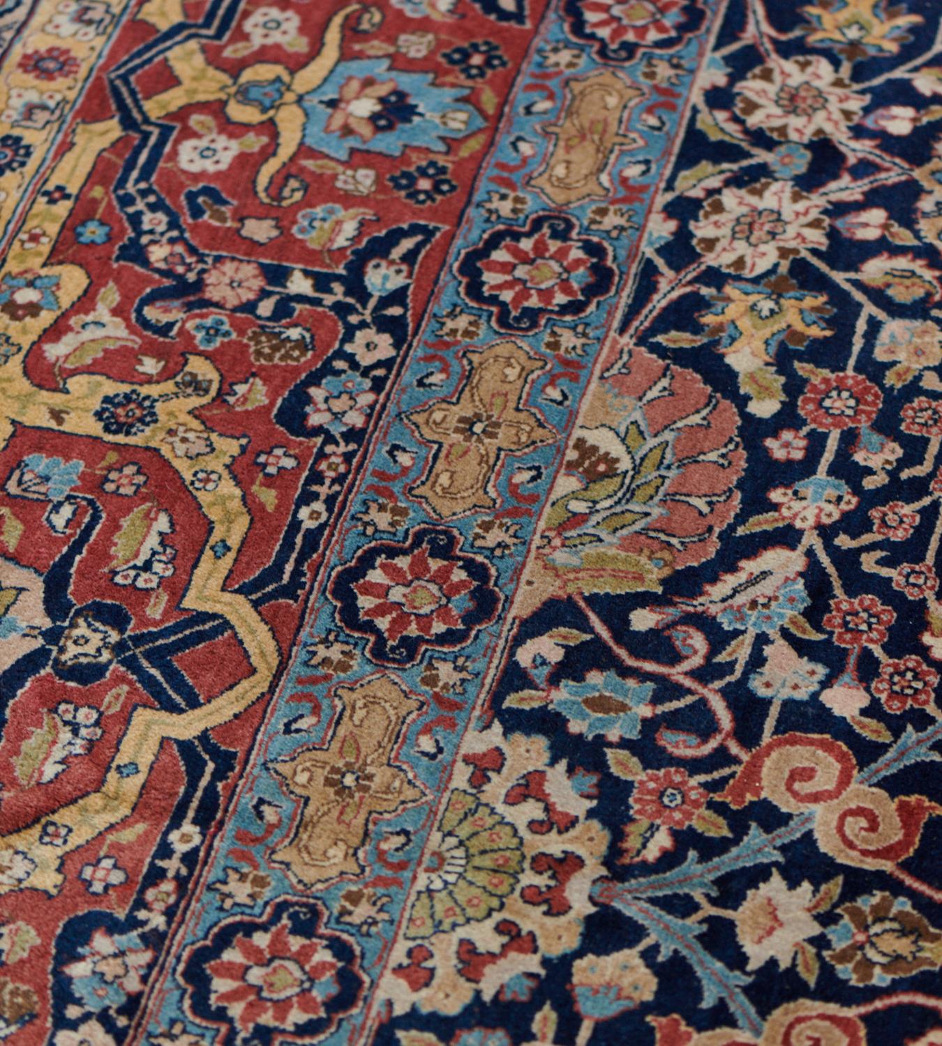 This antique, circa 1910, Persian Tabriz rug has an indigo-blue field with an overall design of polychrome palmette vine surrounded by dense floral sprays and split palmettes, in a broad shaded red border of angular golden-yellow and indigo-blue