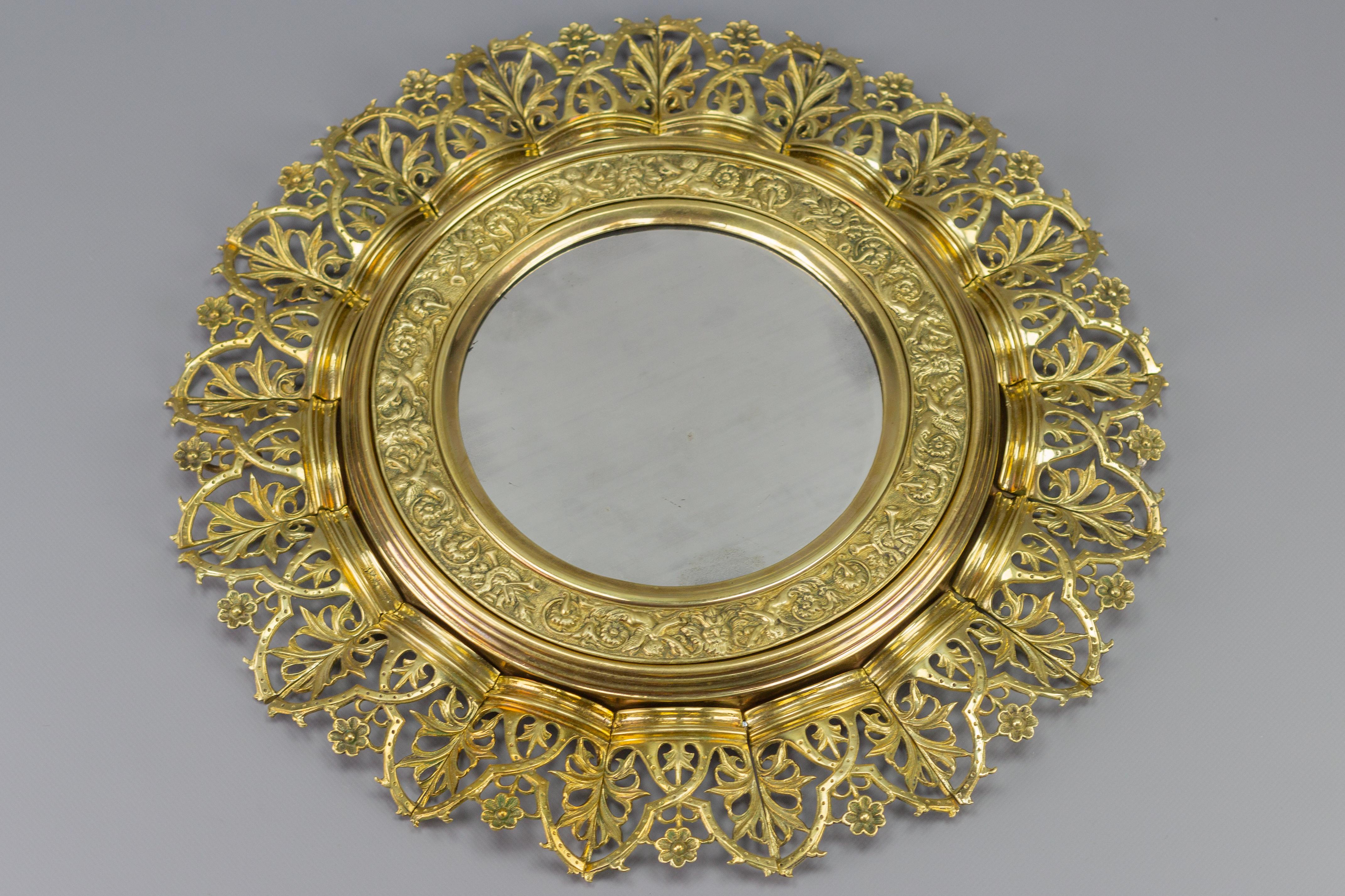 A stunning circular wall mirror in a sunburst shape. This antique mirror features an embossed brass and bronze frame that is richly decorated with beautiful neoclassical details such as putti, flower, and leaf motifs. Crown-like elements in the