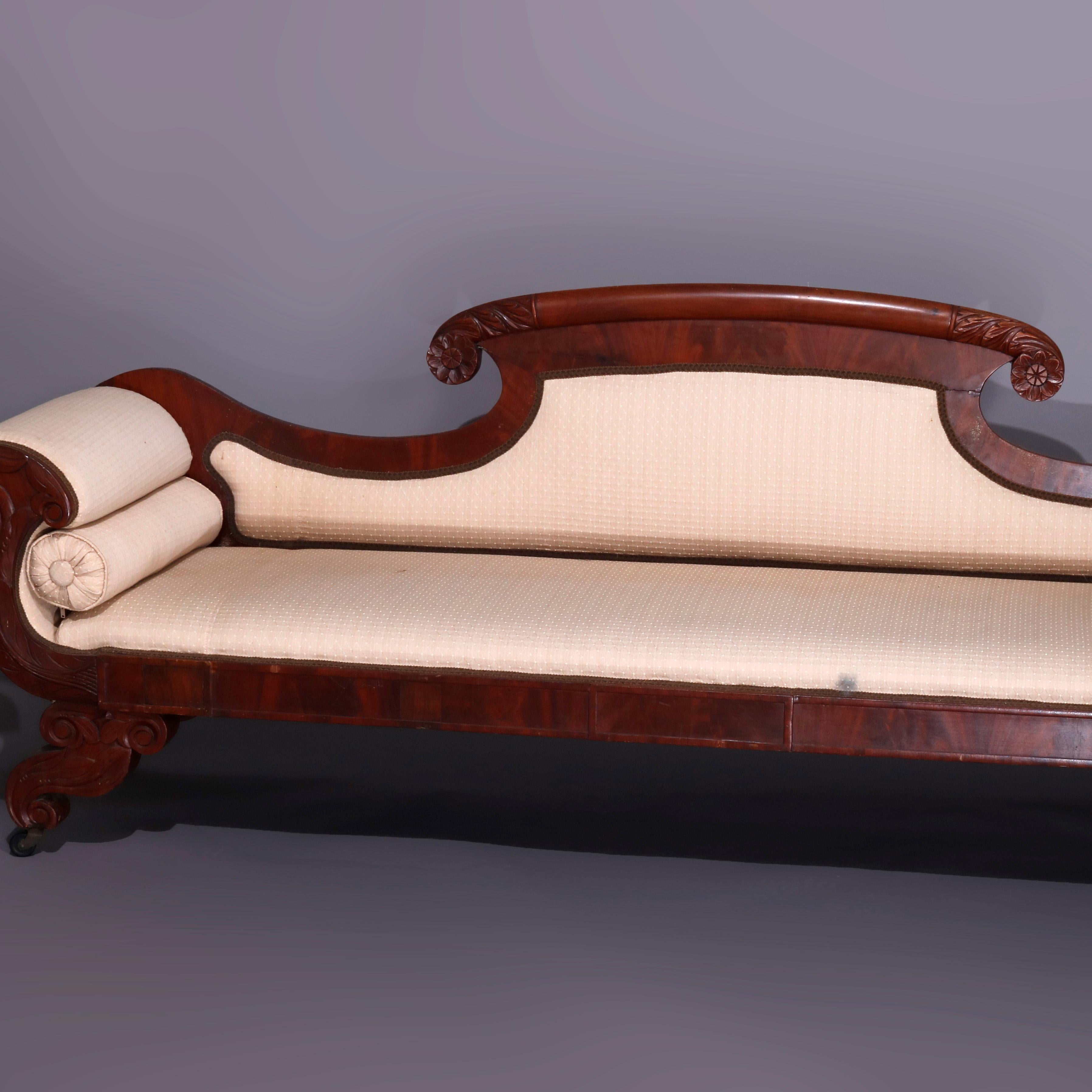 An antique Classical American Empire flame mahogany sofa offers shaped back with curved scroll form foliate carved crest terminating in rosettes and surmounting upholstered back and seat with stylized scroll form arms having foliate carved frame,