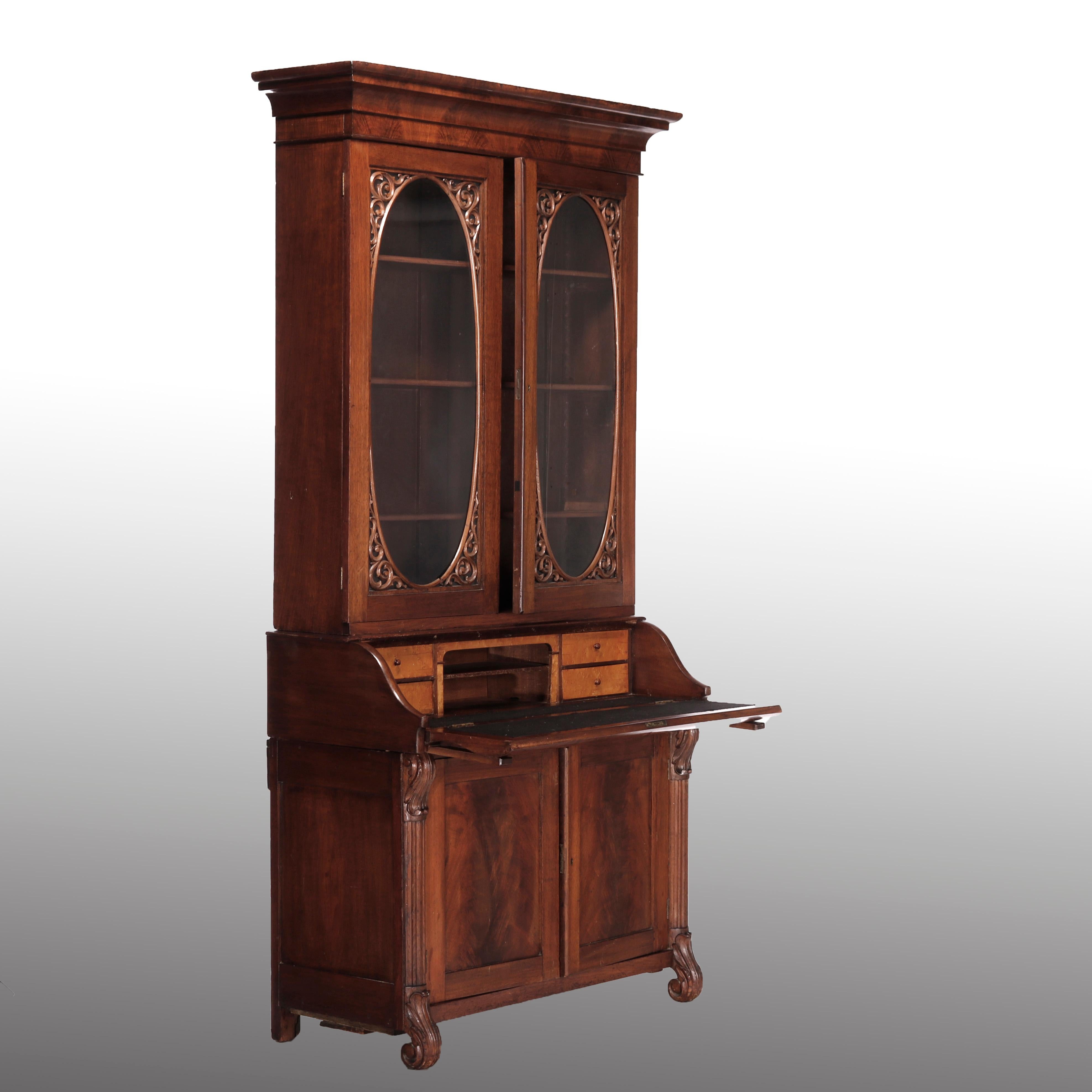 An antique Classical Greco American Empire secretary offers flame mahogany construction with upper having bookcase with double glass doors each with carved scrolled foliate filigree corbels and opening to a shelved interior, over lower case having