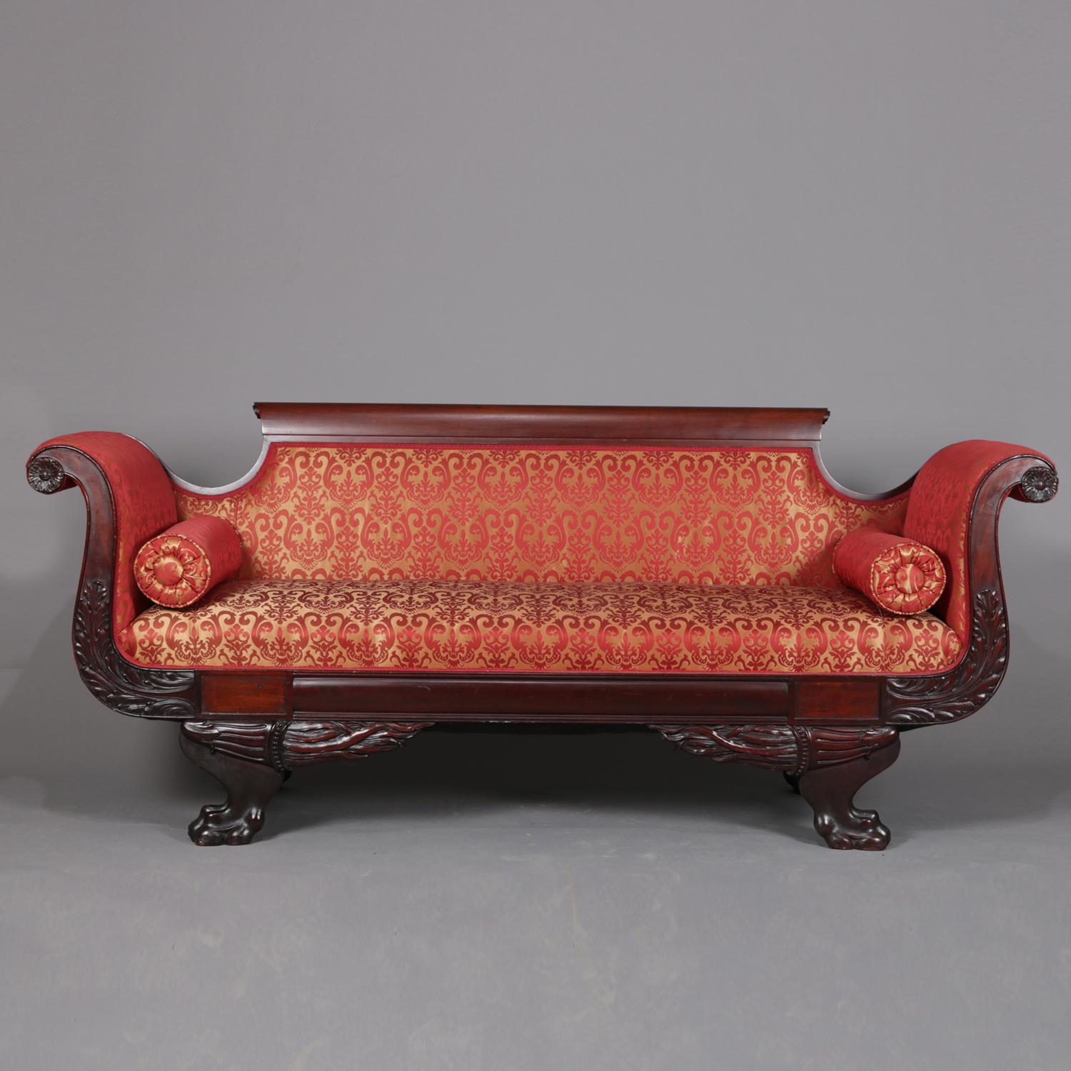 An antique classical American Empire upholstered sofa features flame mahogany frame with scroll form arms having carved acanthus decoration and raised on four carved paw feet flanking a carved foliate skirt, newer upholstery, circa