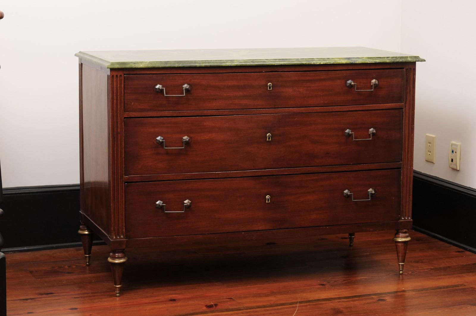An antique classical-style chest. A green marble top rests on the mahogany case that features 3 drawers. Fluted columns on the sides of the case taper into pointed feet.