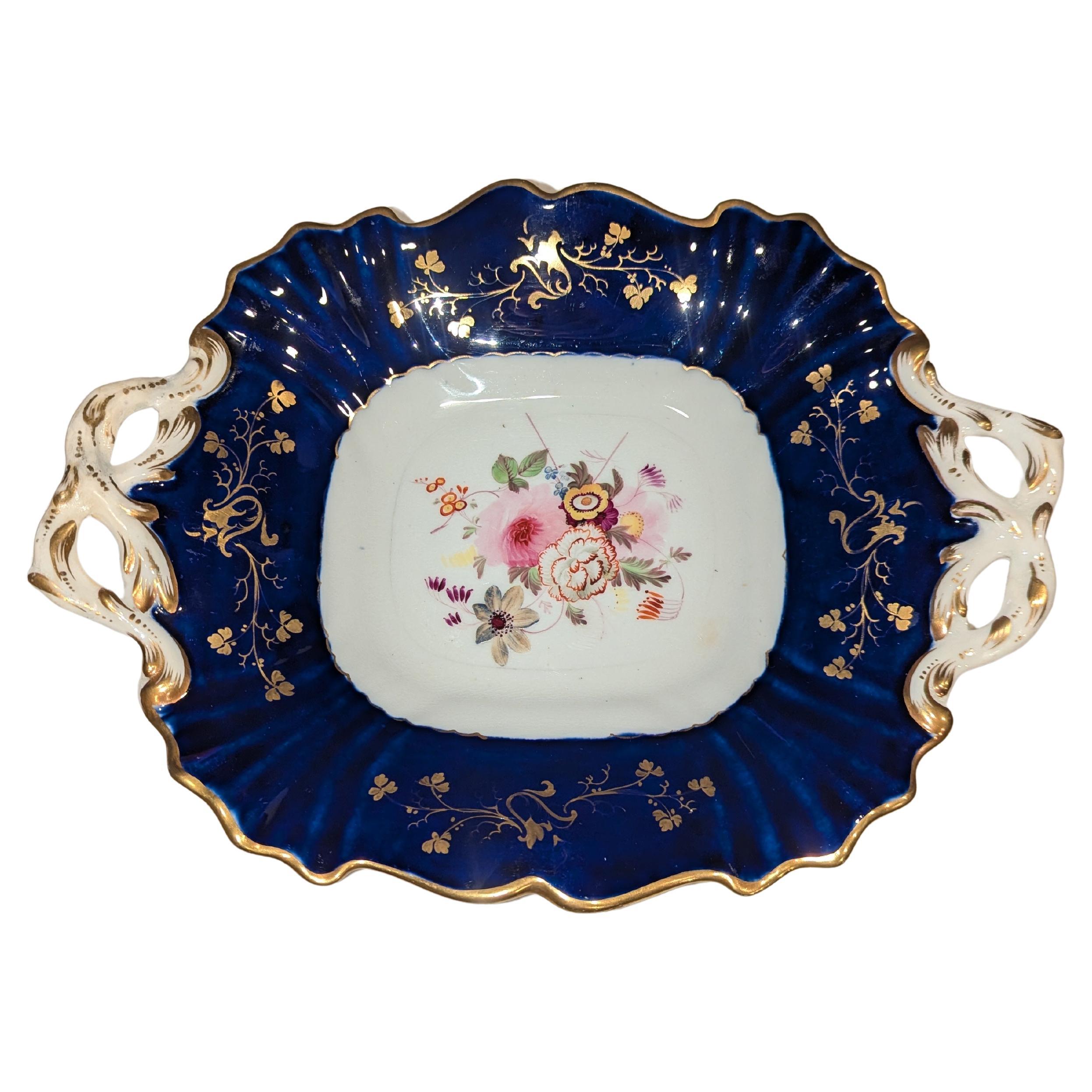 Esta impresionante bandeja antigua con asa y motivo floral dorado y azul cobalto de Coalport es un notable ejemplo de la artesanía en porcelana inglesa del siglo XIX. Elegantemente diseñada, la bandeja presenta un fondo azul cobalto intenso que
