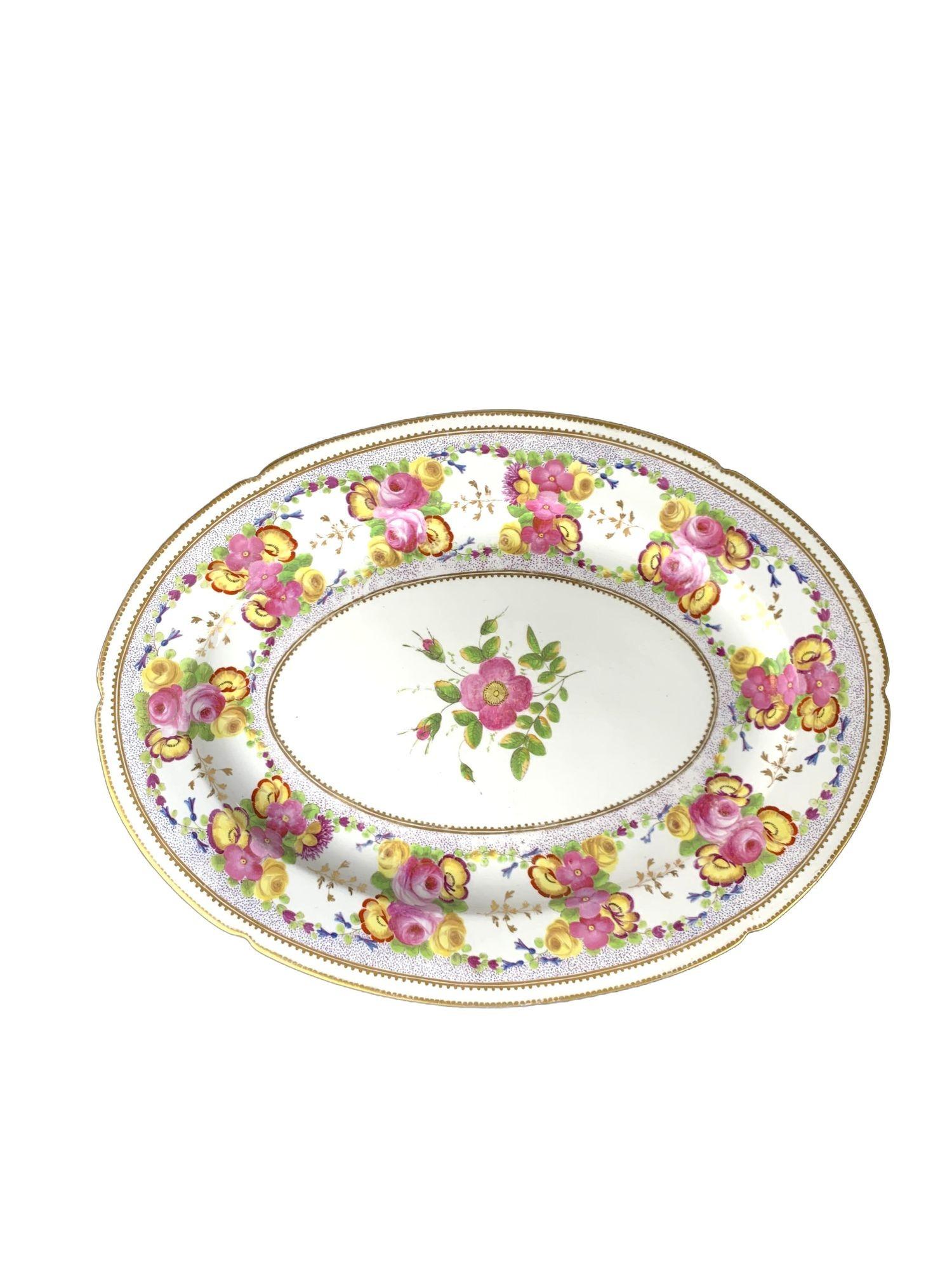 Esta antigua fuente de porcelana fue fabricada por Coalport en Inglaterra hacia 1825.
Está pintada a mano con vivos motivos florales en tonos rosa, amarillo y morado, centrados en un medallón ovalado y repetidos en paneles a lo largo del borde.
Las