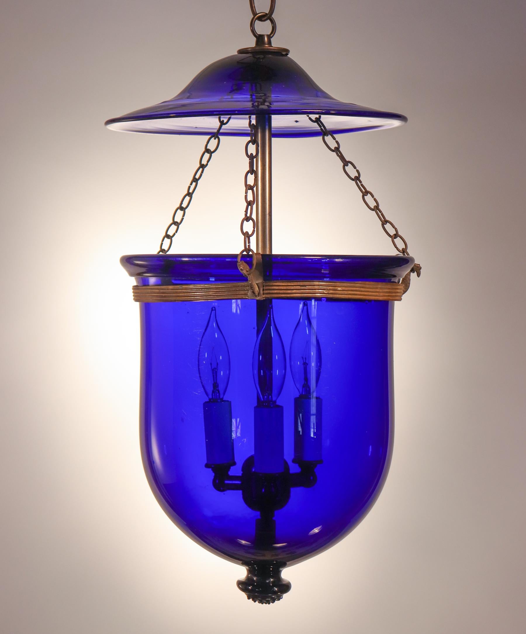 A striking cobalt blue hand blown glass bell jar lantern with its original smoke bell and rolled brass band, circa 1890. Originally for candlelight, this antique pendant has been newly electrified and casts a warm light from its three-bulb