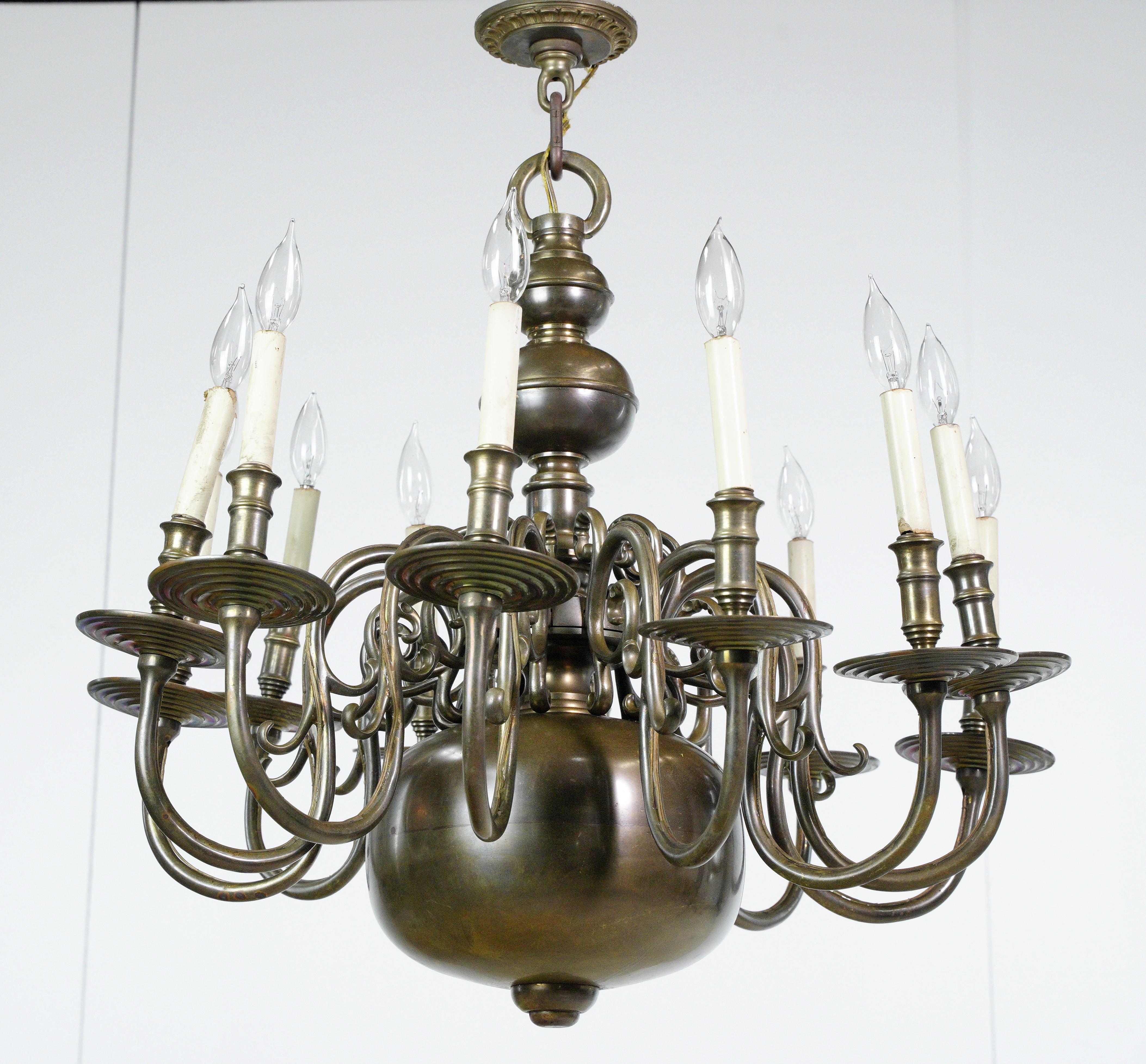Antique Colonial twelve arm bronze chandelier. Crafted with curled arms and a regal presence, this exquisite piece adds a touch of antique opulence to any room. This is in good antique condition with some surface wear. The price includes restoration