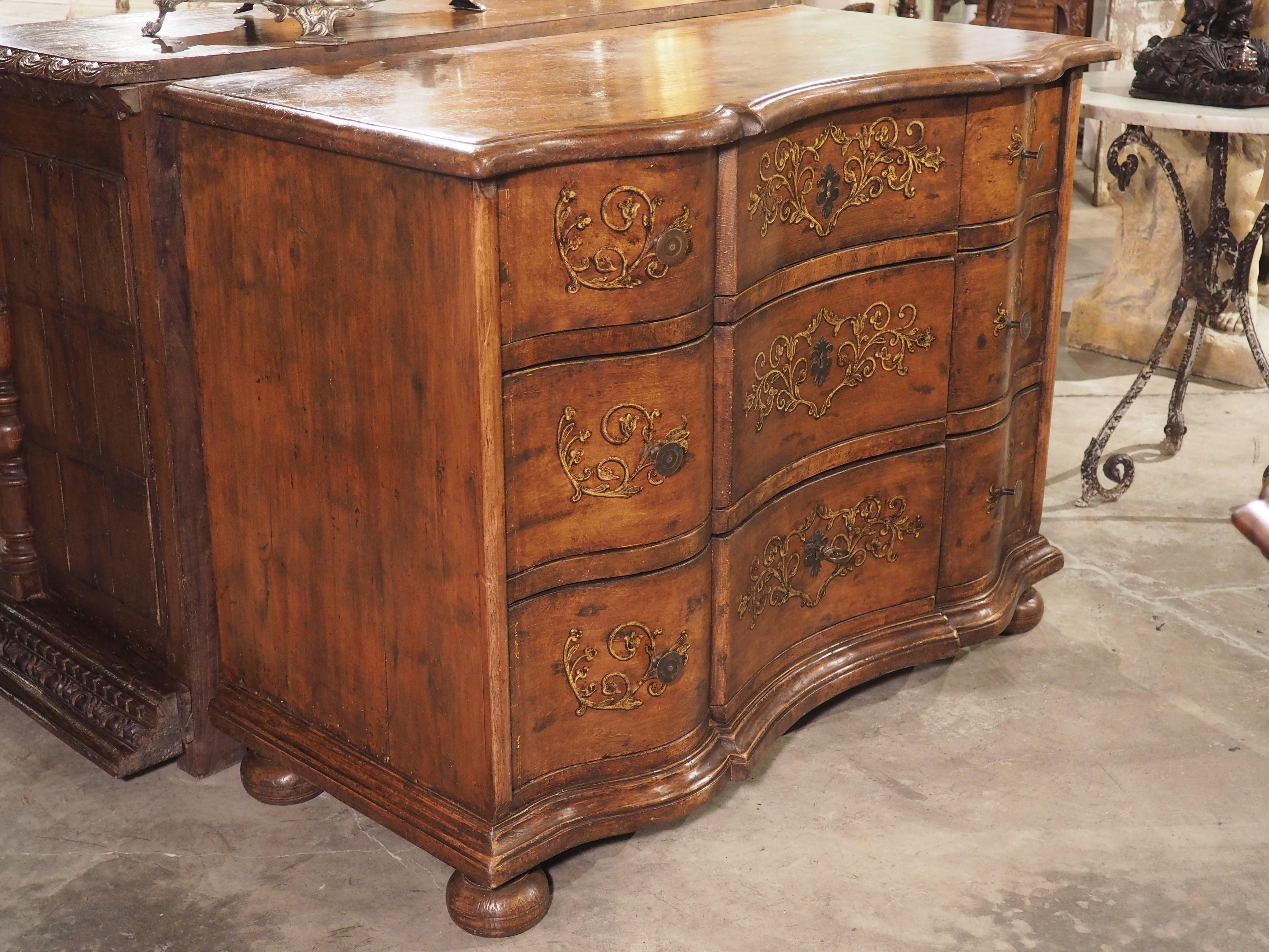 Hand-carved in Northern Italy during the 1800’s, this antique commode has lovely gilt painted drawer fronts. The hand-painted embellishments feature sprawling floral rinceaux and C-scrolls with a golden-brown hue. The airy and curvaceous nature of