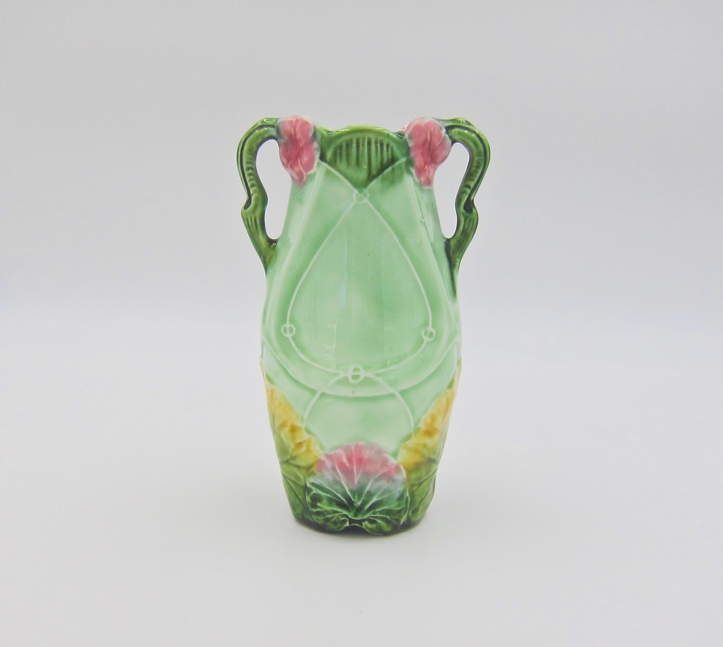 An antique majolica vase in the European Art Nouveau style, dating to the late 19th century. The double-handled vessel is decorated with molded blossoms and leaves glazed in vibrant pink, yellow, and green against a ground of pale green. The