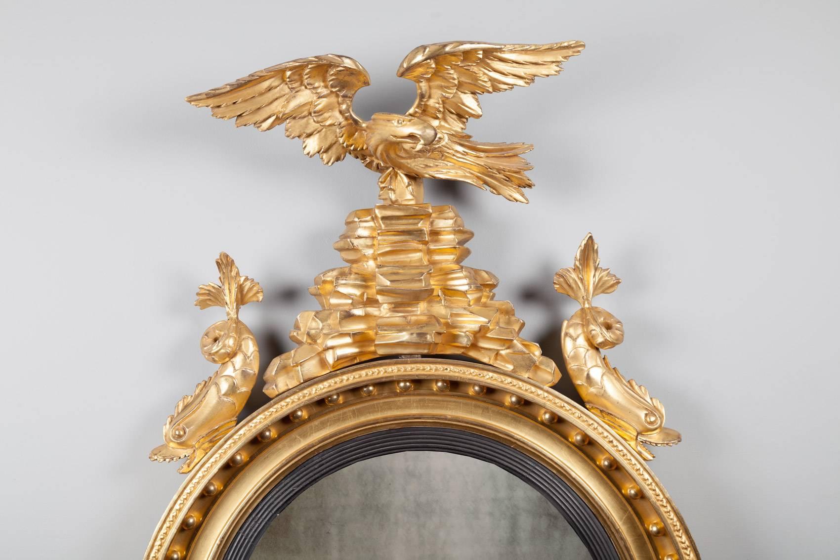 A fine quality antique English Regency giltwood convex mirror. The original convex mercury mirror is set in a reeded ebonized frame, which is surrounded by musket ball ornament. An eagle perched on rock-work is flanked by two stylized dolphins. The