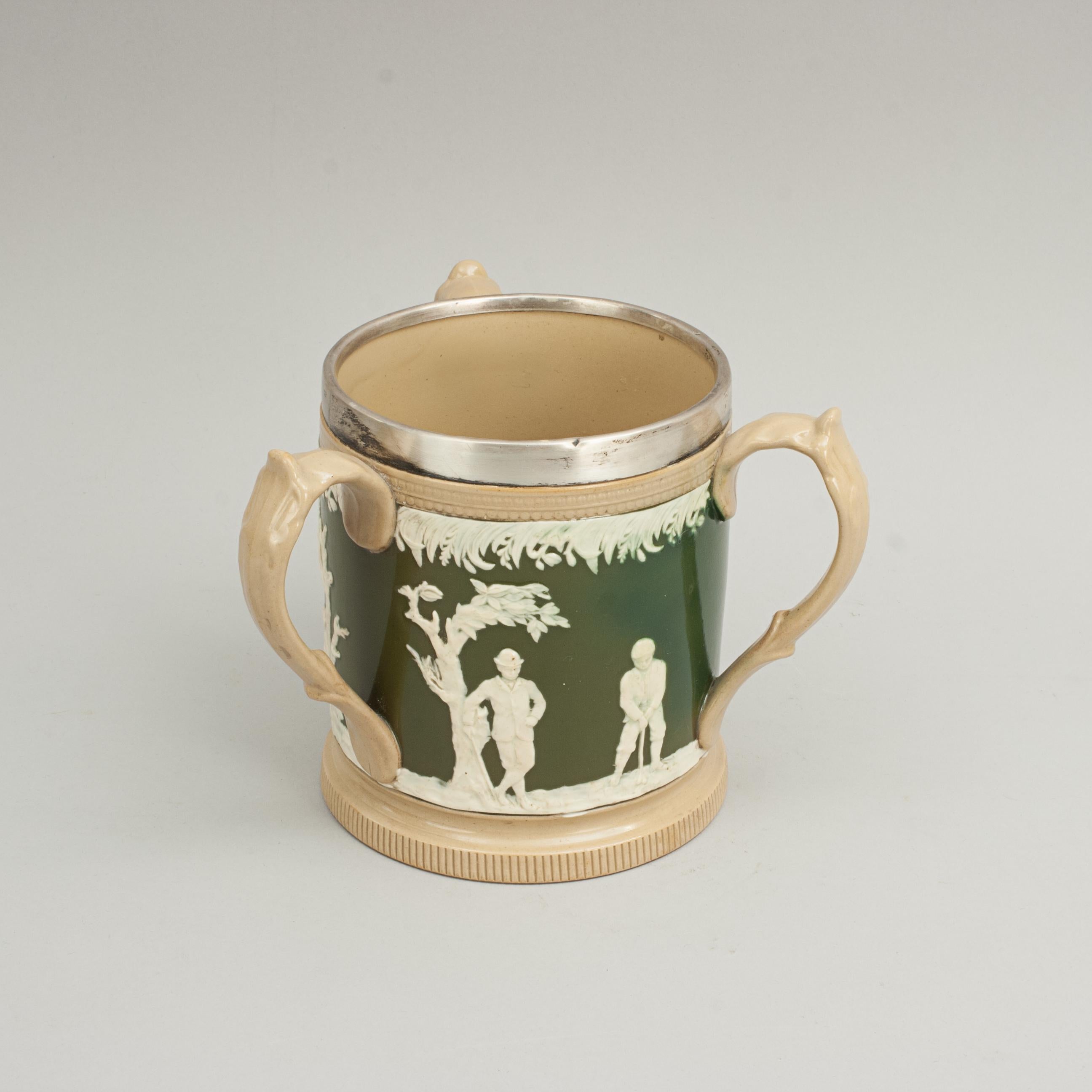 Three handled golf loving cup, Tankard.
A fine Copeland Spode tyg with silver rim, three handles and three golf scenes. This wonderful golf tankard decorated with golfers in white relief on olive green and beige background, the silver rim