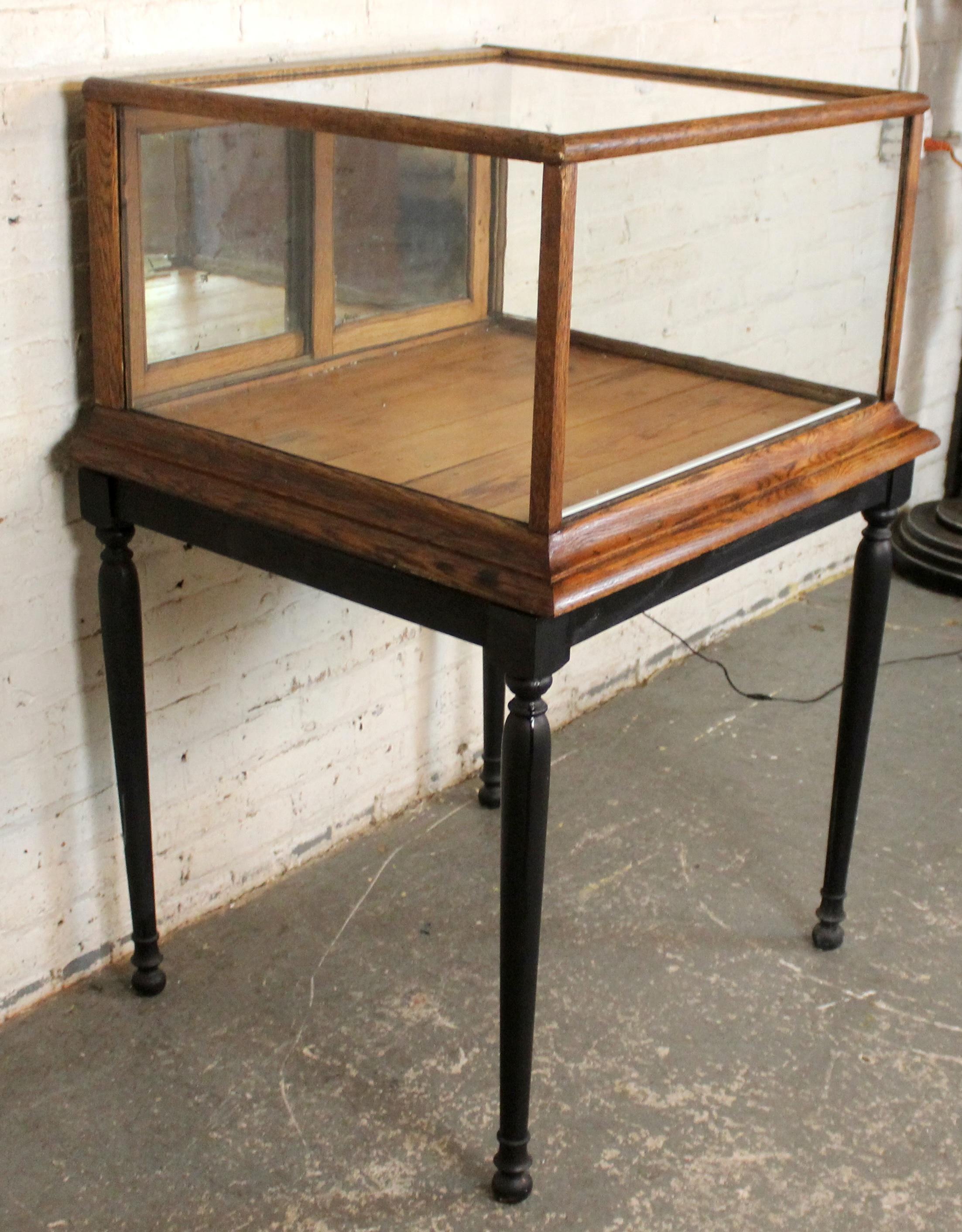 A charming antique countertop display cabinet featuring a warm wood frame, clear glass panels, and a sliding rear door for access. Raised on tall, turned black legs, it has a light, elevated presence that makes it ideal for showcasing small