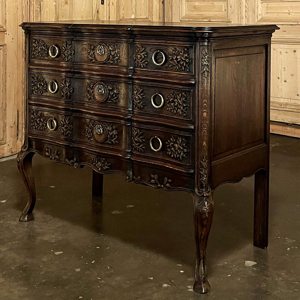 Antique Country French Regence commode features an exquisitely contoured facade festooned with elaborate wildflower, foliate and laurel wreath motifs. Scrolled bordering on the contoured apron combines with the rounded and carved cornerposts to
