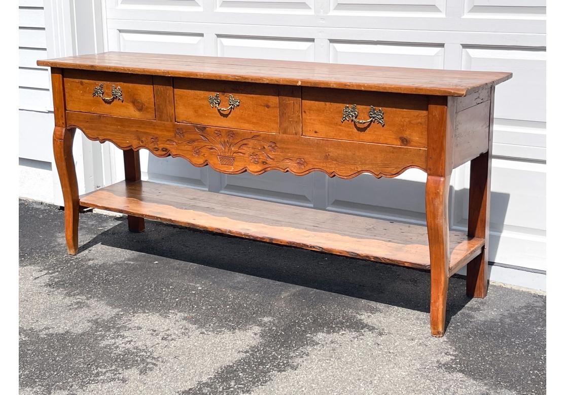 Serveur rustique français ancien avec dessus en planches bien usé et construction à chevilles, trois tiroirs avec poignées en laiton bien moulées. La table présente un tablier festonné et sculpté avec un rebord décoratif et un motif de plante en pot