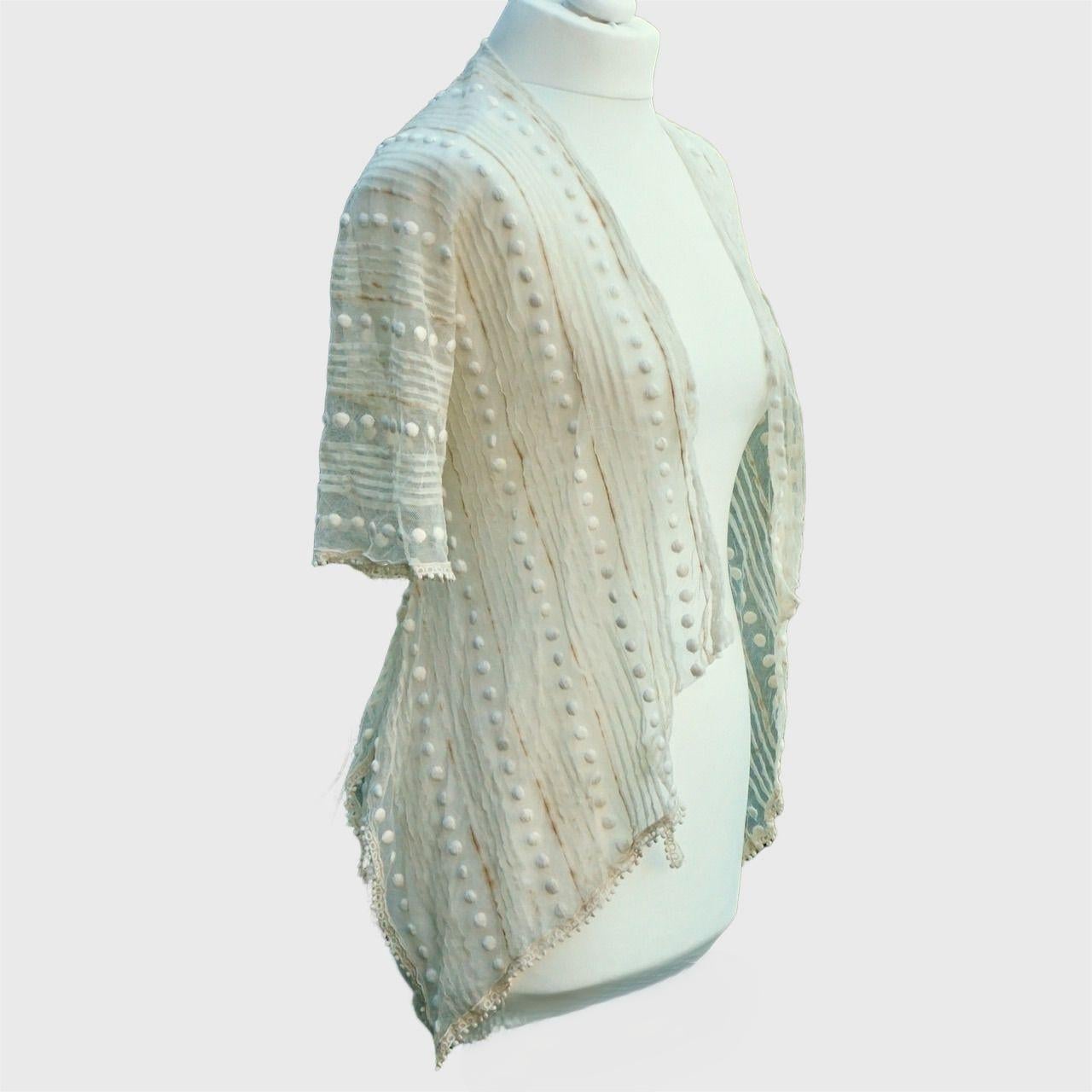 Magnifique veste en dentelle de filet crème antique, avec un motif de plis et de cercles en tissu. Il présente un ourlet en forme d'écharpe, terminé par des bordures fantaisie. Une partie des bordures est manquante à une extrémité. Les manches sont
