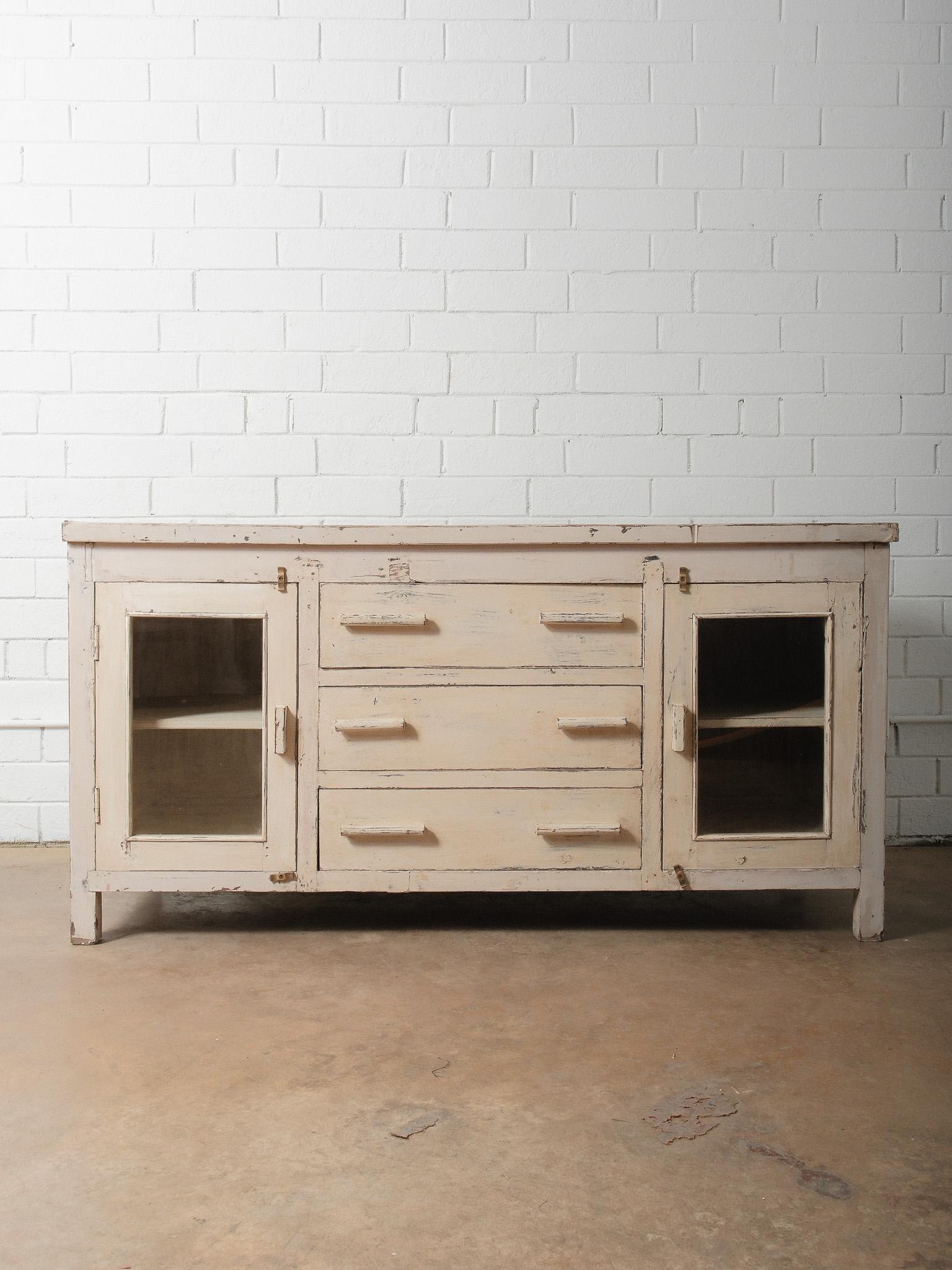 Buffet ancien en bois peint en crème, riche en caractère, avec une peinture joliment vieillie, des portes vitrées et trois larges tiroirs centraux. Cette pièce basse et substantielle présente des décennies d'usure authentique, avec une peinture en