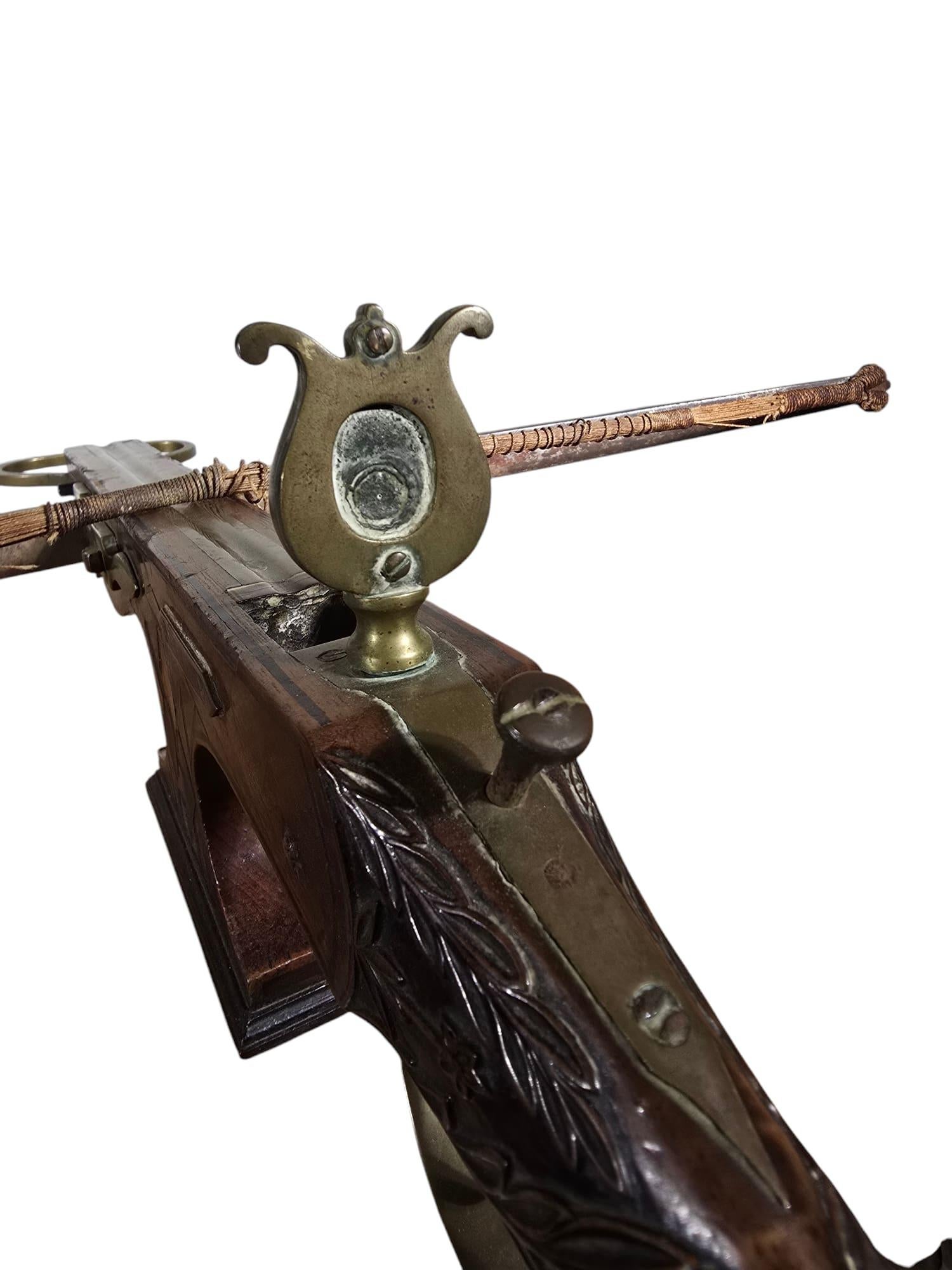 This is an antique crossbow from the 19th century or earlier, masterfully crafted with fruitwood, steel, and bronze. The stock features an elegant design, adorned with intricate carvings and fine marquetry, showcasing exceptional craftsmanship. The