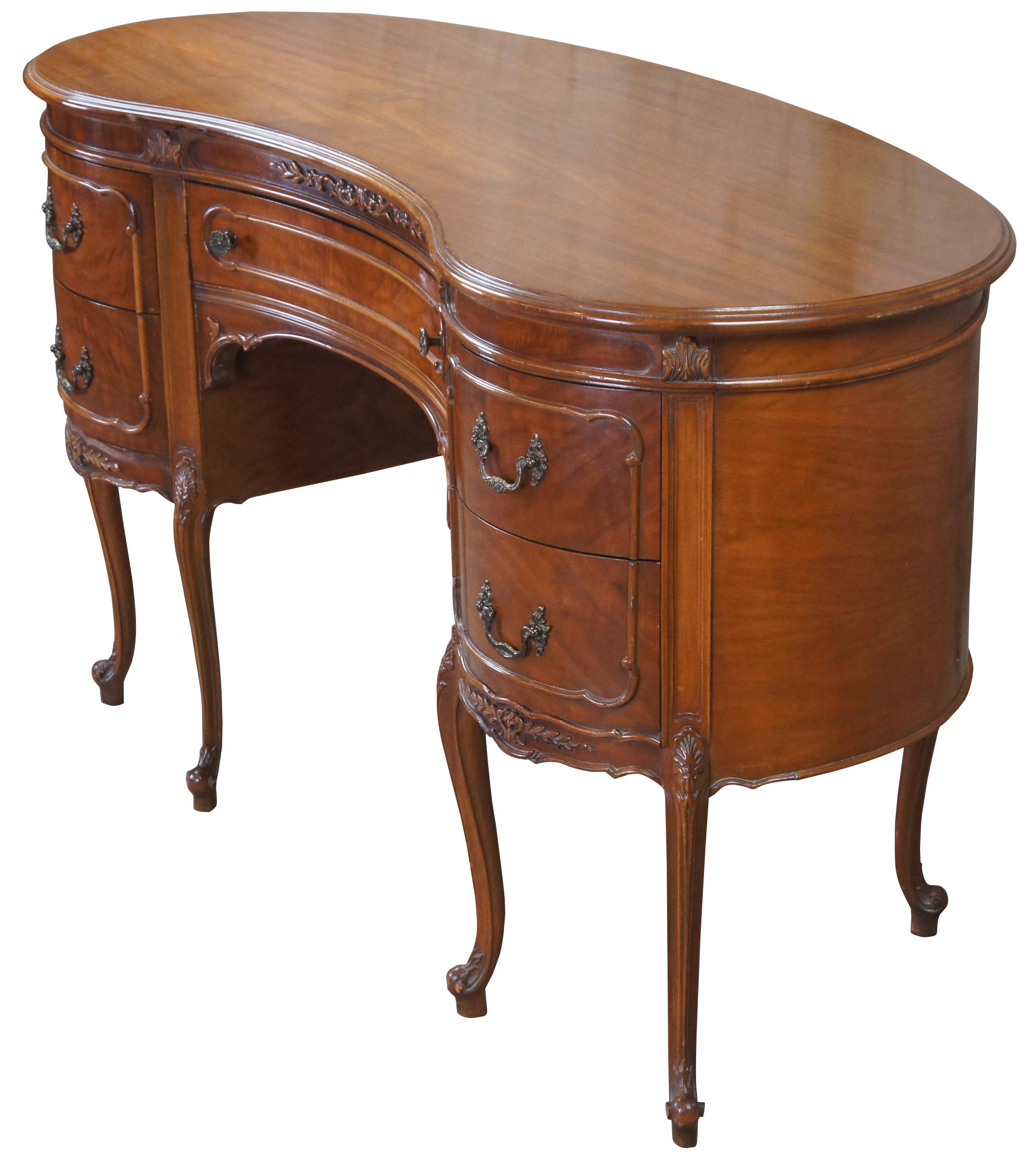 1940s kidney bean or crescent shaped writing desk. Made from walnut with crotch veneer along the front. Features French styling with carved foliate accents and five drawers supported by six cabriole legs over scrolled feet. Includes glass tops.