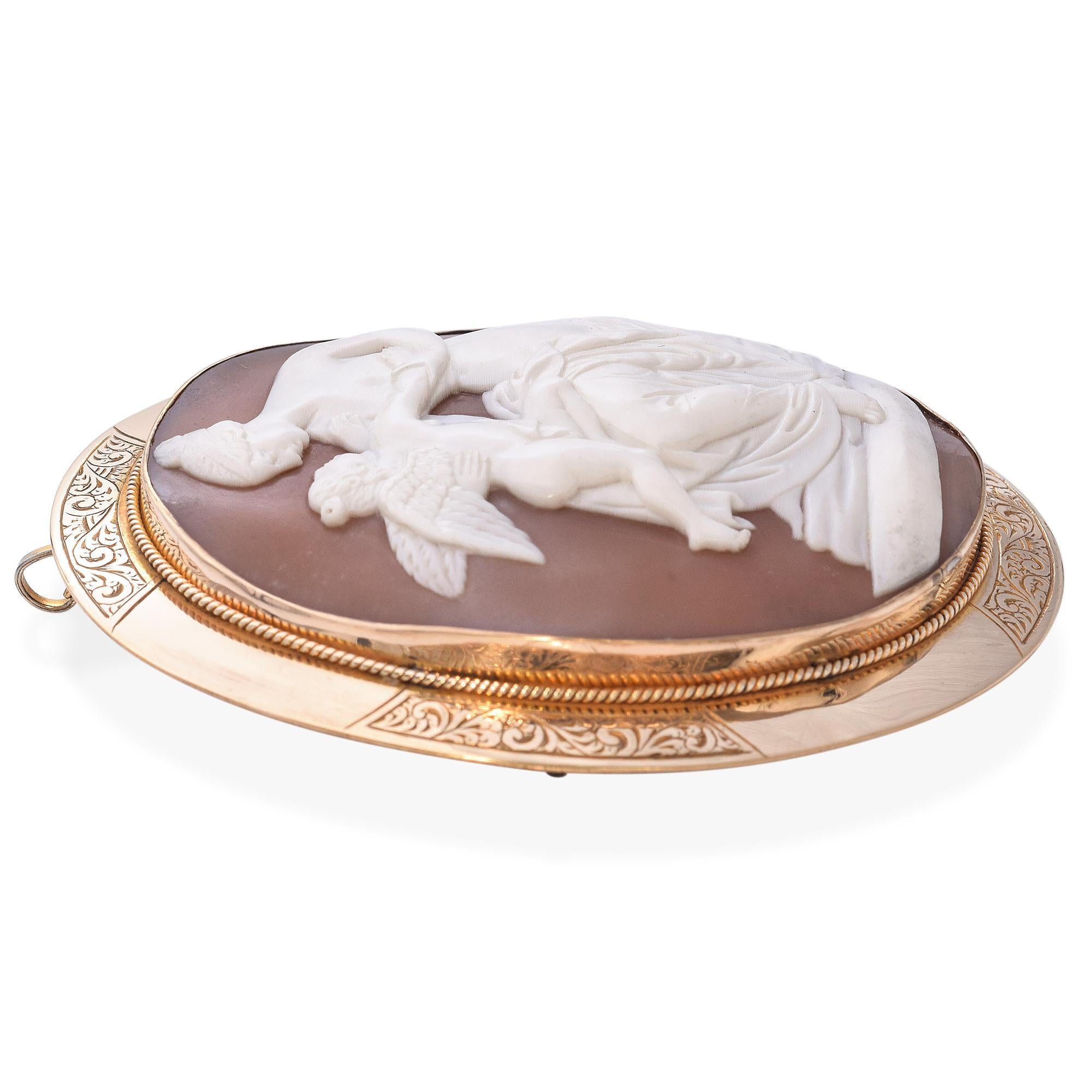 Diese zarte und fein geschnitzte antike Muschelkamee-Brosche fängt einen intimen mythologischen Moment ein: Amor, der Gott der Liebe, wiegt sich sanft auf dem Schoß seiner Mutter - wahrscheinlich Venus (Aphrodite), der Göttin der Schönheit und