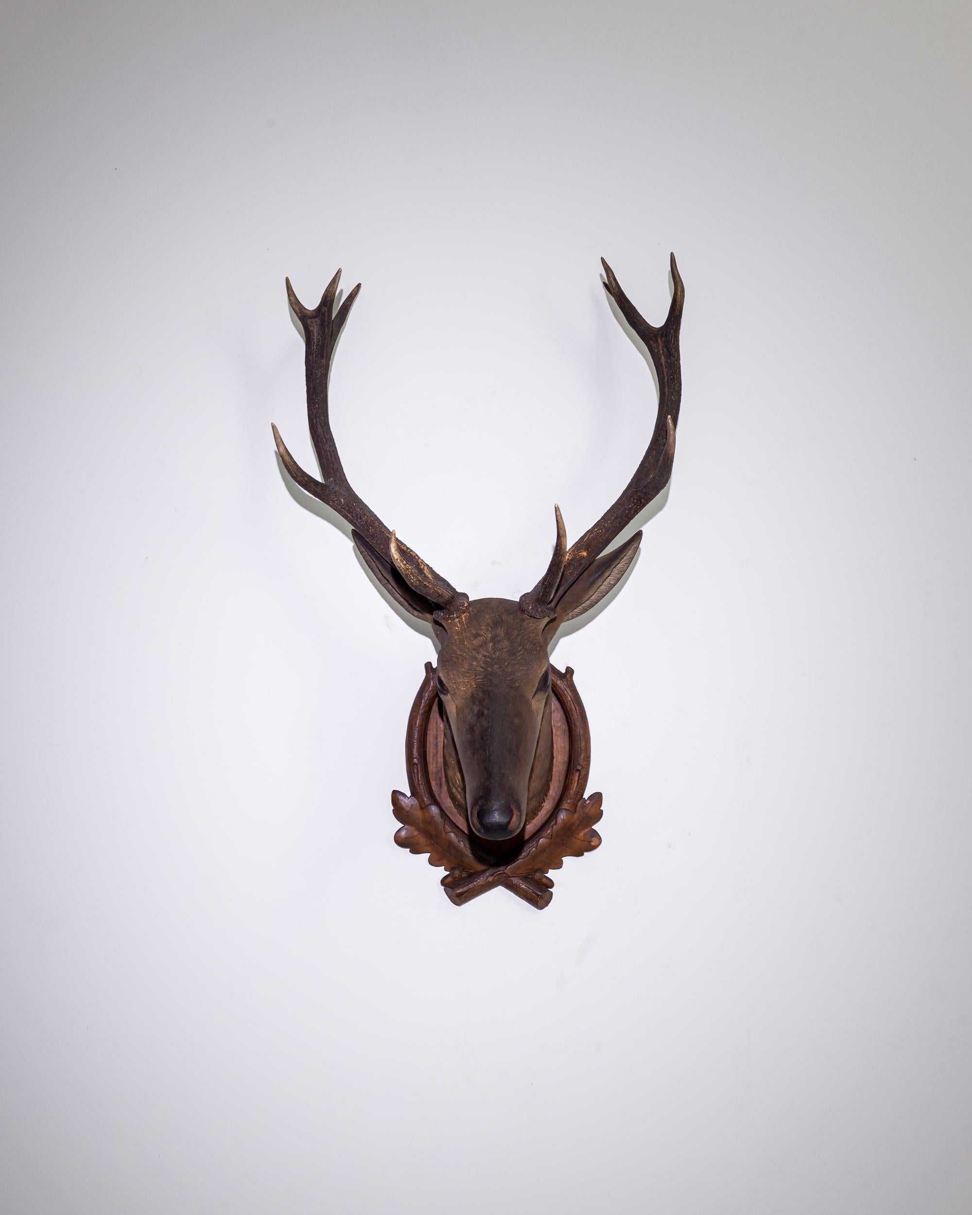 This wooden stag’s head decoration evokes the rustic backdrop of a mountain hunting lodge. Made in Czechia in the 20th century, a pair of genuine antlers are mounted atop a carved wooden stag’s head. The dark finish of the wooden frame lends the
