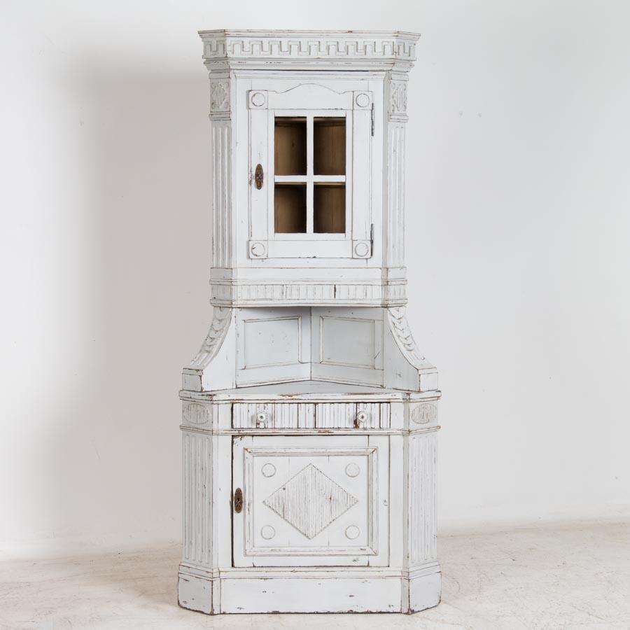 The light gray paint is softly distressed exposing the natural pine beneath, enhancing the crown molding and carved details of this Classic Danish corner cupboard. Made in three sections, the door below feature diamond elements often seen in