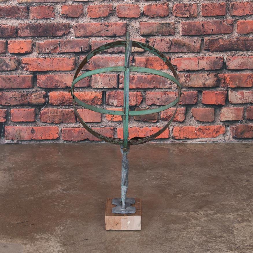 This vintage globe shaped garden ornament, called an armillary, is made up of metal bands pierced by an arrow and supported by a figural mounting bracket. The remnants of old green paint on the outside and an aged brass patina inside (with number