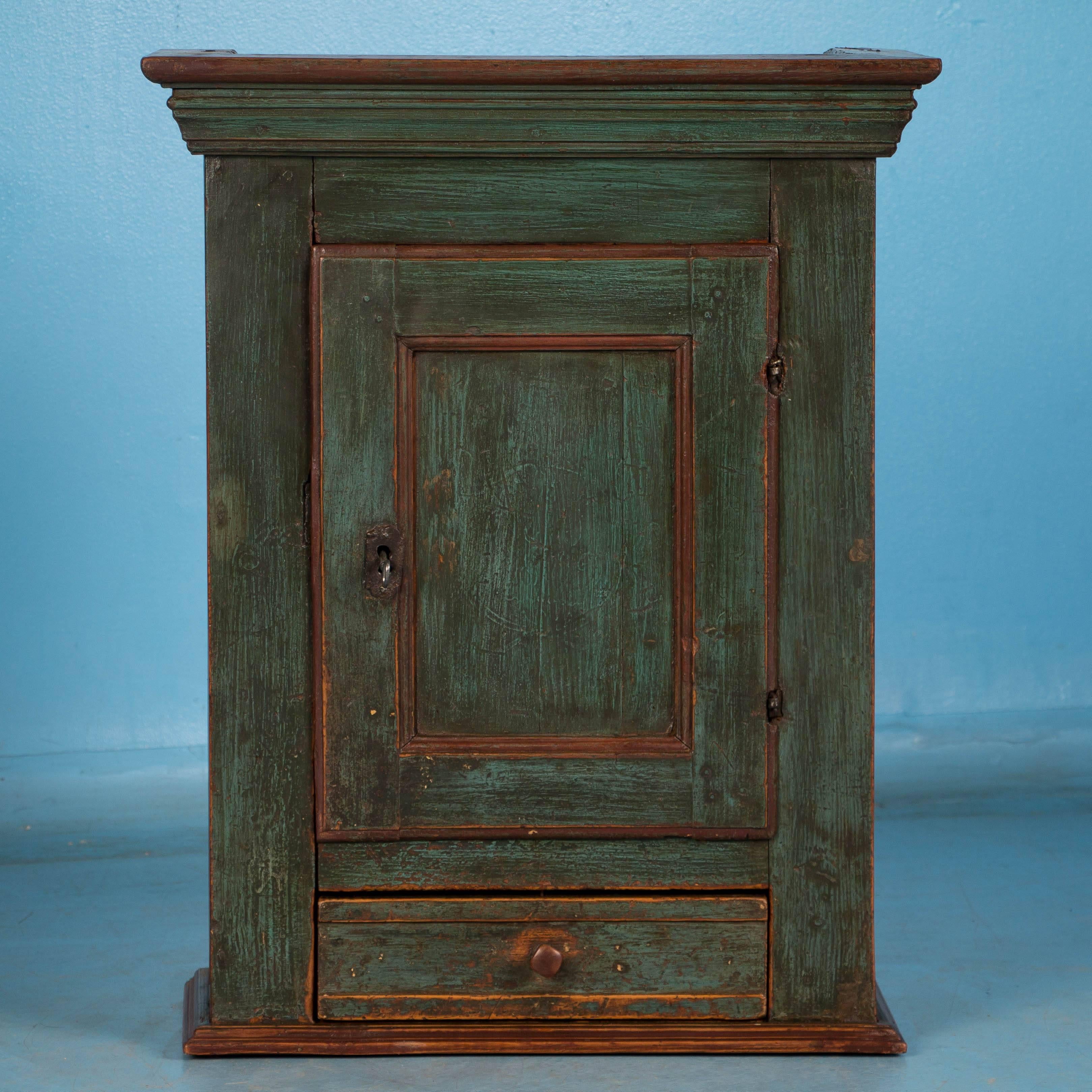 This charming hanging cupboard from Denmark is exceptional due to the aged patina of the original green paint over the pine case. Take note of the two shelves inside and the unique pin that locks the drawer. The cabinet has been professionally