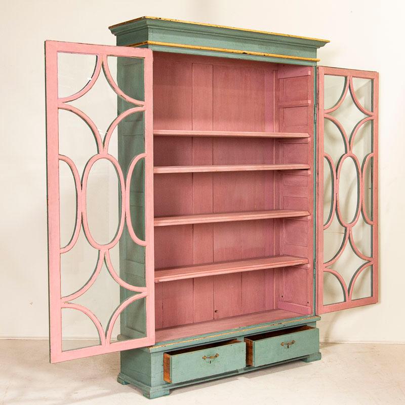 This elegant bookcase has graceful decorative trim over glass doors, showing off any collection stored inside. The attractive exterior blue paint is accented with gold trim, while the interior pink creates a romantic contrast. The two lower drawers