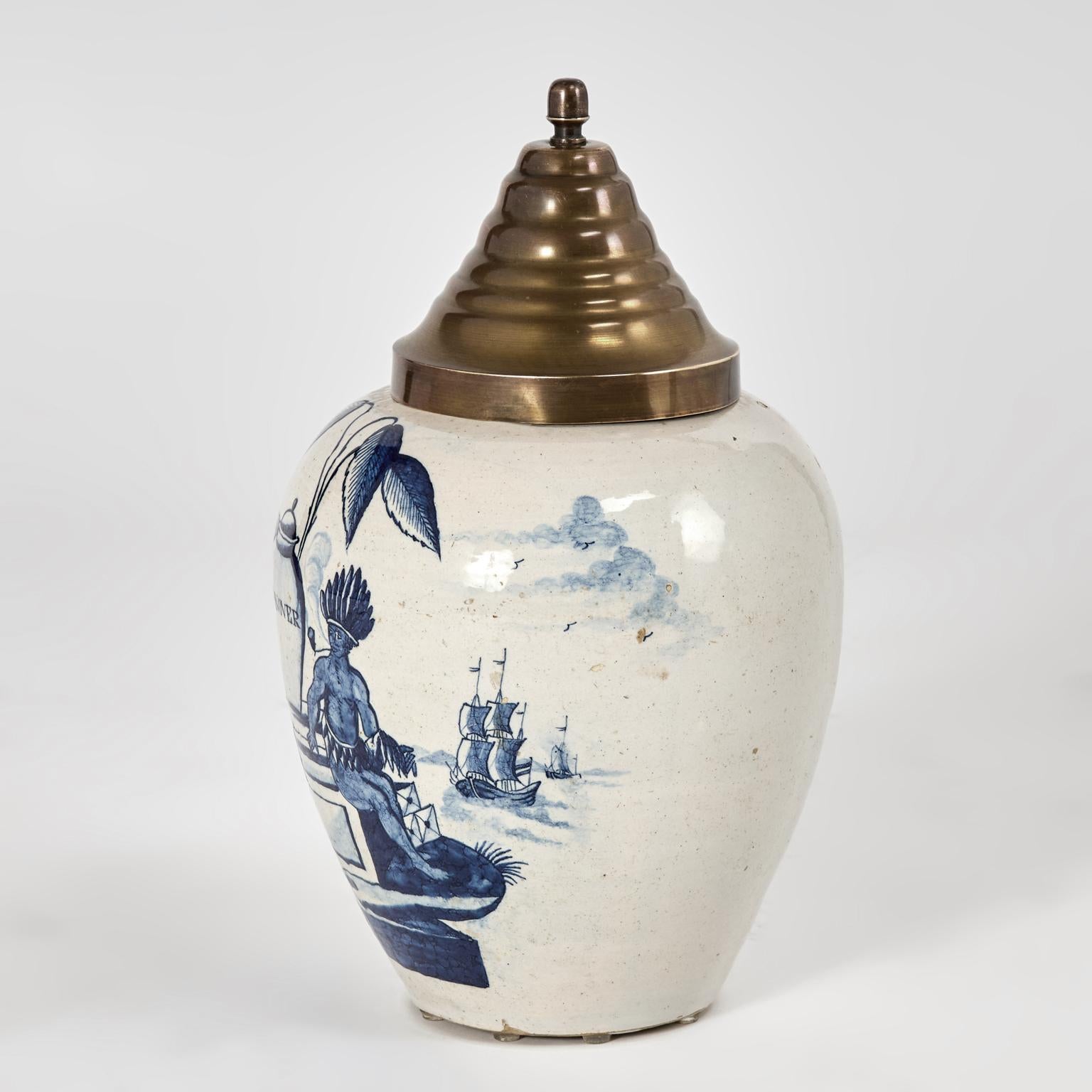 Un barattolo per tabacco con coperchio in terracotta delft del XVIII secolo, decorato a mano con piante di tabacco, urne, una nave e un nativo americano in un affascinante blu su uno sfondo color crema. Il coperchio è in ottone e il vaso è siglato