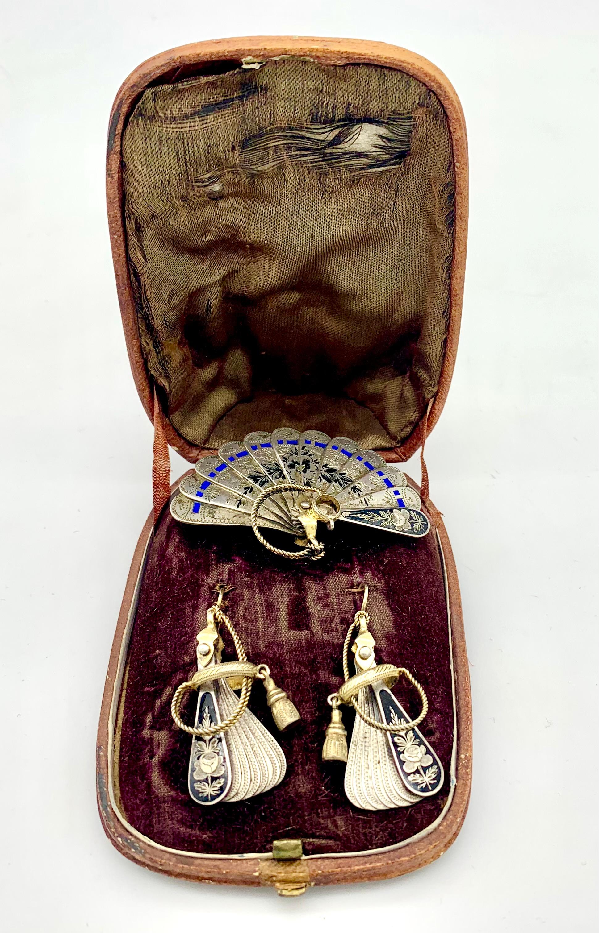 This unusual demi parure in its original box comprises a pair of dangling earrings and a brooch in the shape of fans.
The fans are made out of silver and are finely engraved with a flower motifs and decoratet with blue and black enamel-