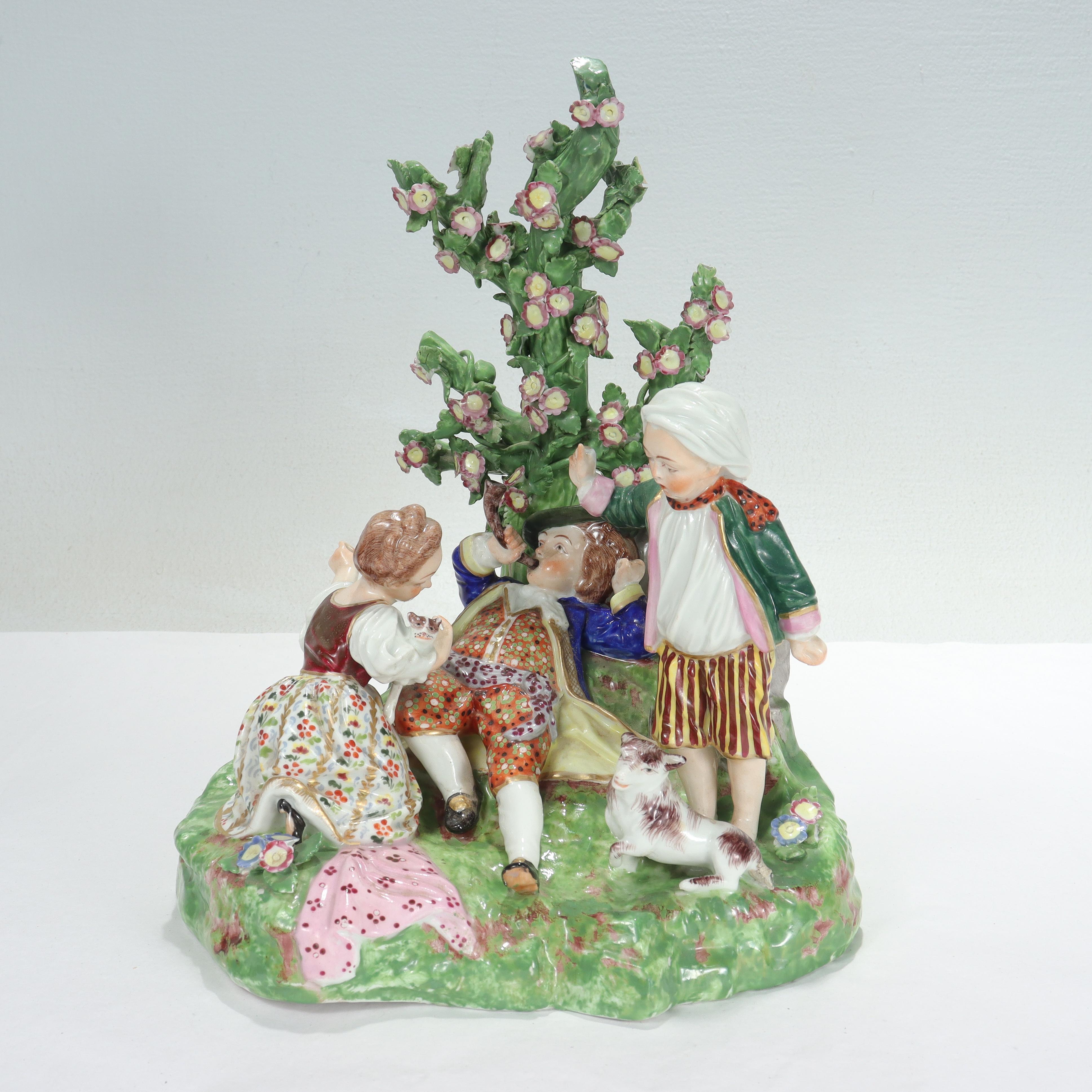 A fine antique English porcelain figurine.

By the Derby Porcelain Works.

With 3 children in a garden accompanied by a lamb and a cat. 

The boy is reclining against a tall flowery plant and holding a pipe or scoop in his hand.

Simply a