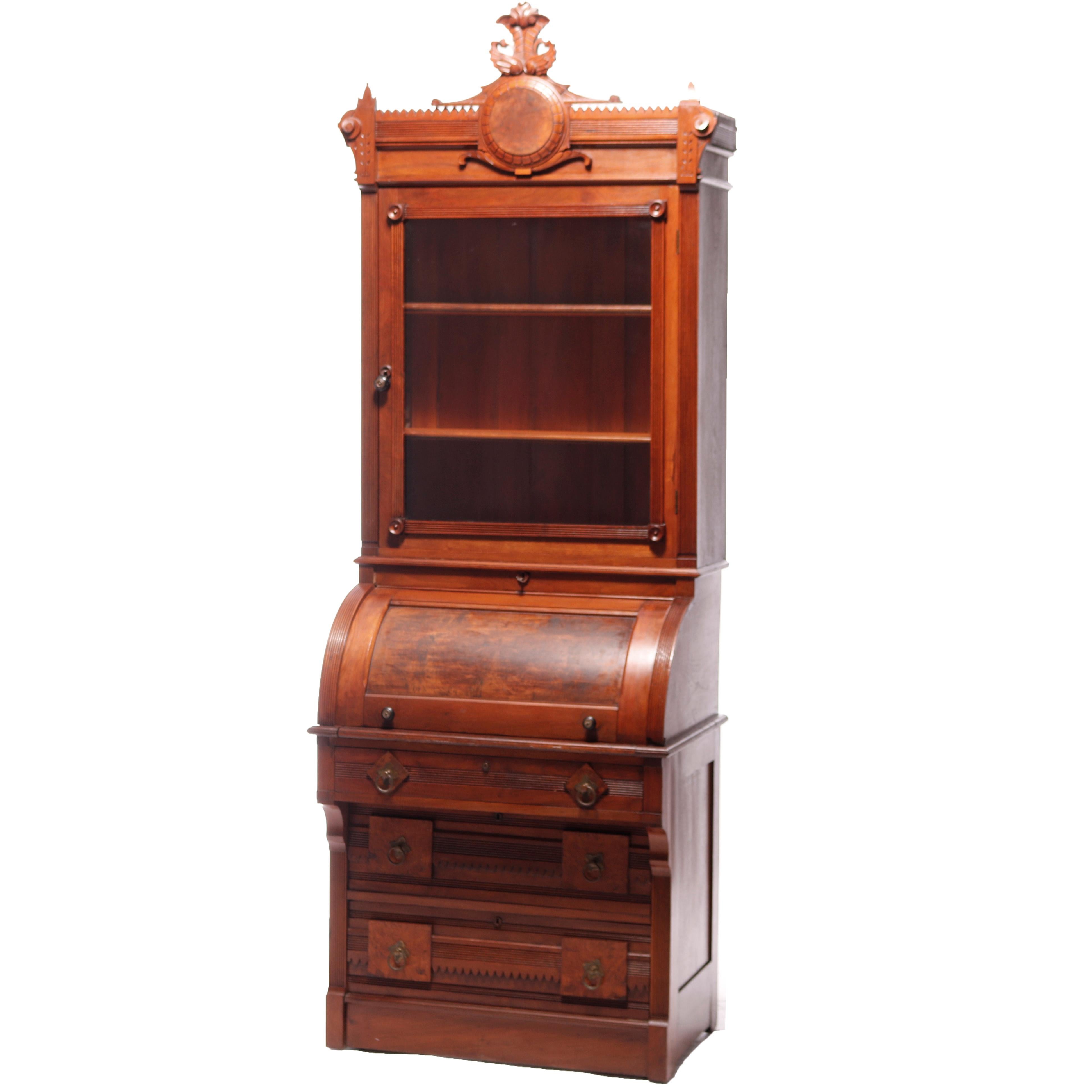 An antique Renaissance Revival diminutive cylinder desk offers walnut and burl construction with upper case having carved cartouche over double glass door bookcase surmounting lower case with cylinder top opening to pull out desk and storage