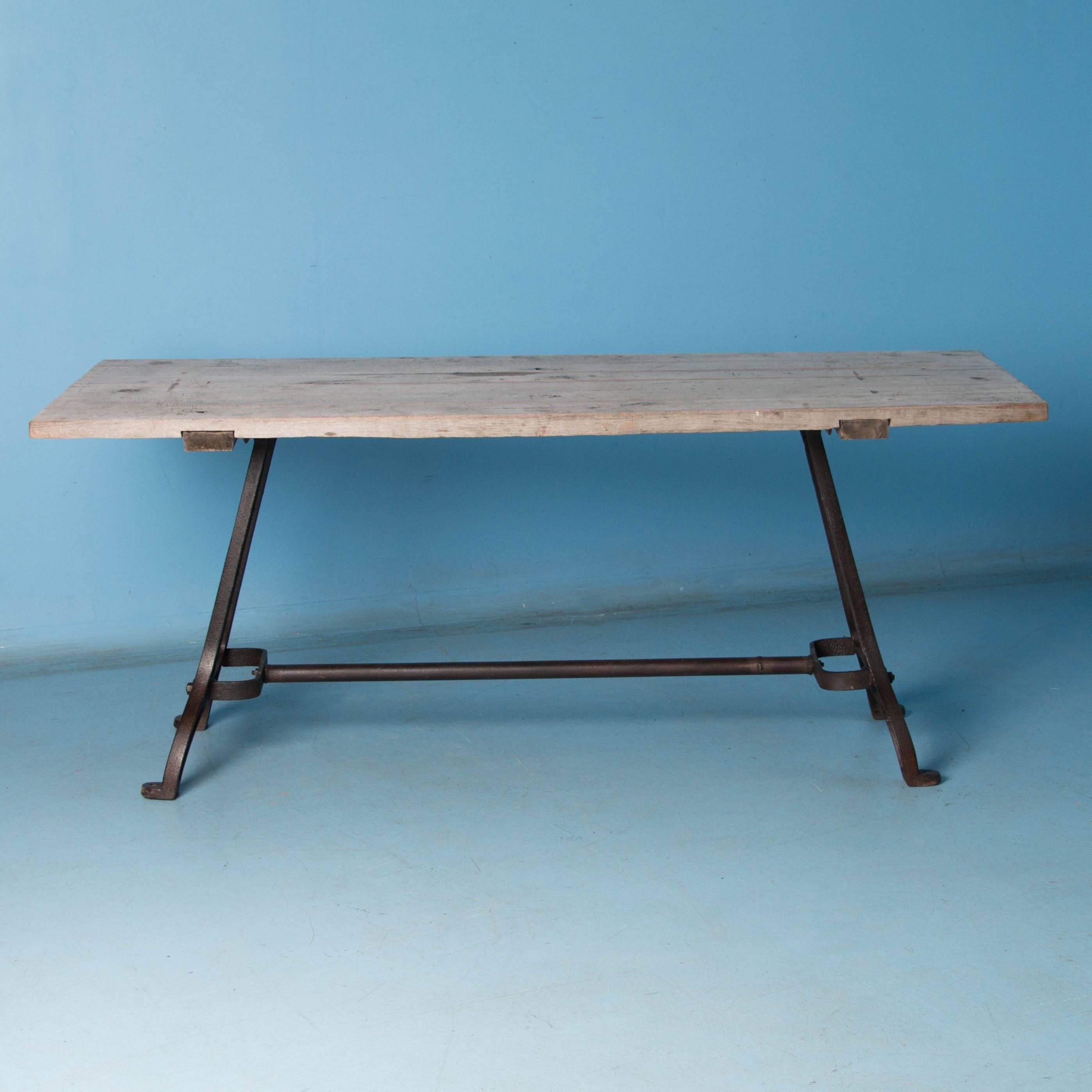 This eastern European dining table with rustic industrial charm, features antique cast iron legs and a hardwood plank top. The distressed silver painted top has the patina of weathered gray wood while the base is a dark gray, almost black, waxed