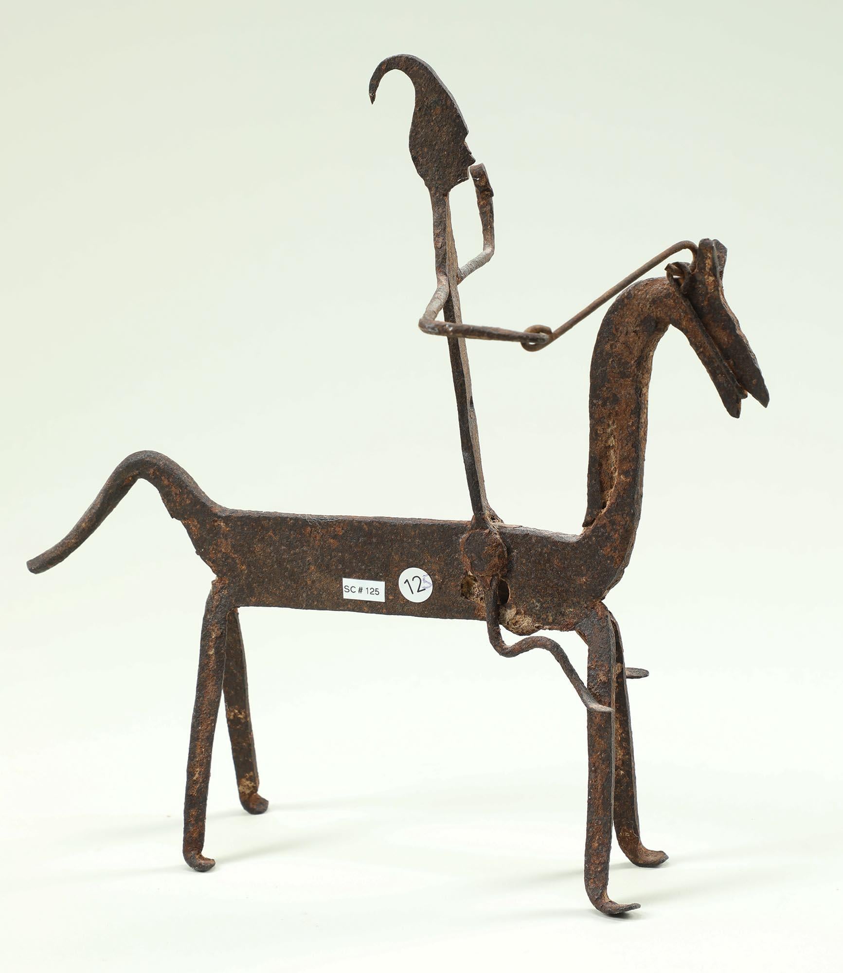 Caballo dogón de hierro forjado con figura.  La figura lleva un sombrero con cresta y tiene una mano en las riendas, la otra curvada en el aire para mantener el equilibrio.
Está en buen estado, adecuado a su edad, uso y soporte. Erosión y óxido por