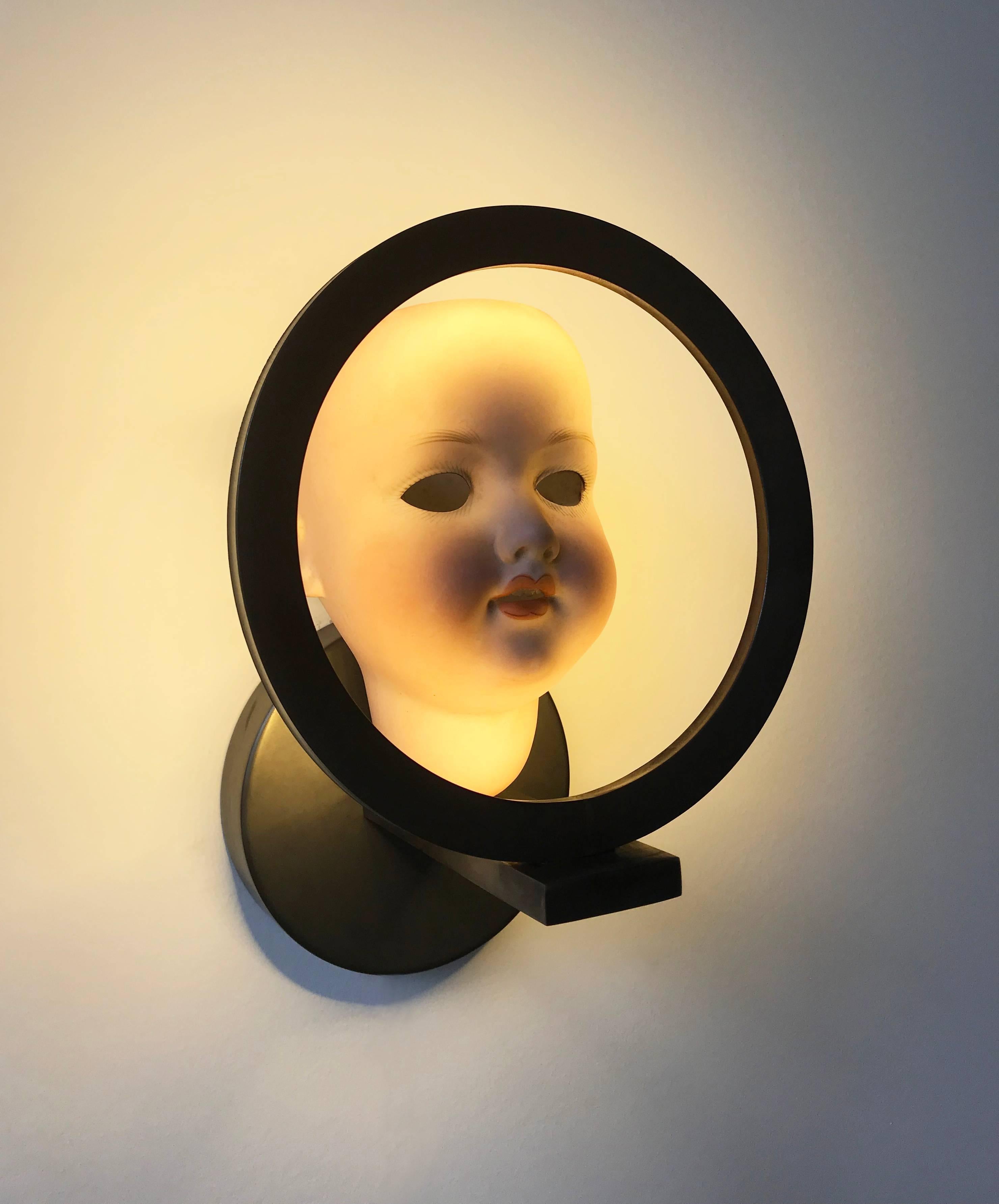 A 'wall light' consisting of an antique ceramic doll head and brass fitting with a dark rustic finish. 
Looking through the eyes, the interior back has been gold-leafed. 
The fitting is handmade in Italy.
The doll heads vary in size, country of