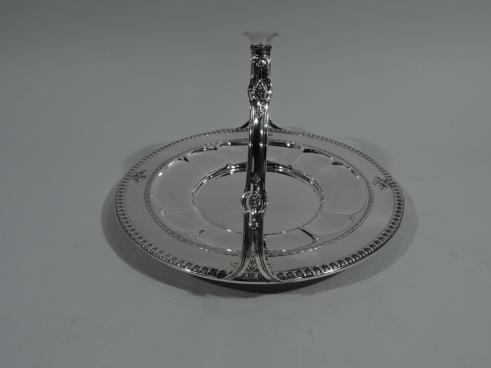 Pretty sterling silver cake plate in Windsor pattern. Made by Dominick & Haff in New York, ca 1915. Circular well. Sides are alternating narrow and wide lobes. Shoulder has chased ribbon-and-flower motif. Bead-and-reel rims. Handle shaped and fixed